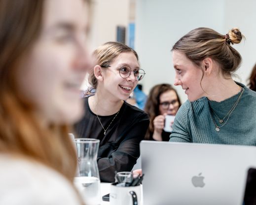 Unsere Kolleginnen Franzi und Fabienne unterhalten sich lachend bei einer Veranstaltung, Laptops und Getränke auf dem Tisch.