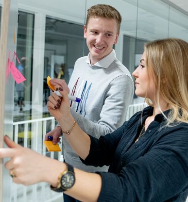 Unsere Kollegen Ole und Alessandra kleben gemeinsam Post-its an eine Glaswand und tauschen sich dabei aus.