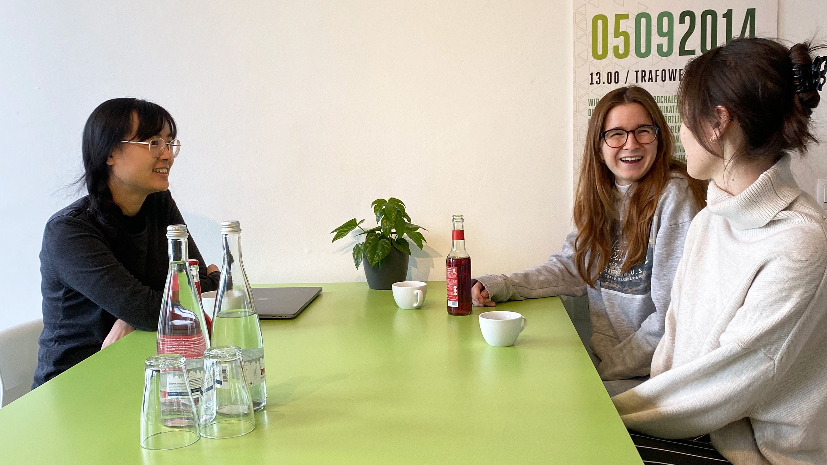 Die ottomisu Mitarbeiterinnen Anita, Ann-Kathrin und Claudia sitzen an einem grünen Tisch im Küchenbereich der Agentur. Claudia spricht.