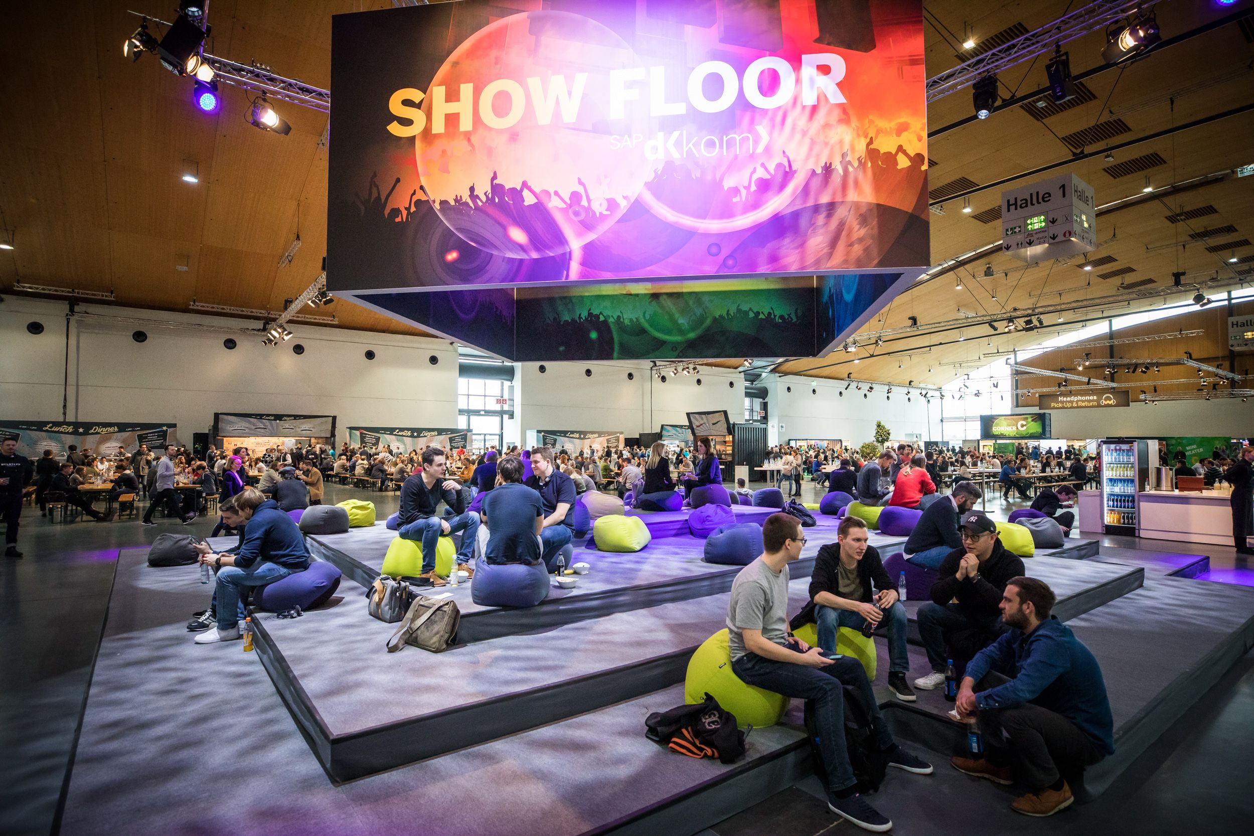 Ein atmosphärisches Bild des Set-Ups der SAP DKOM. Zu sehen ist eine Tribüne, auf der die Besuchende auf Sitzsäcken sitzen und sich miteinander unterhalten. Über ihnen befindet sich ein Bildschirm in Form eines Würfels.