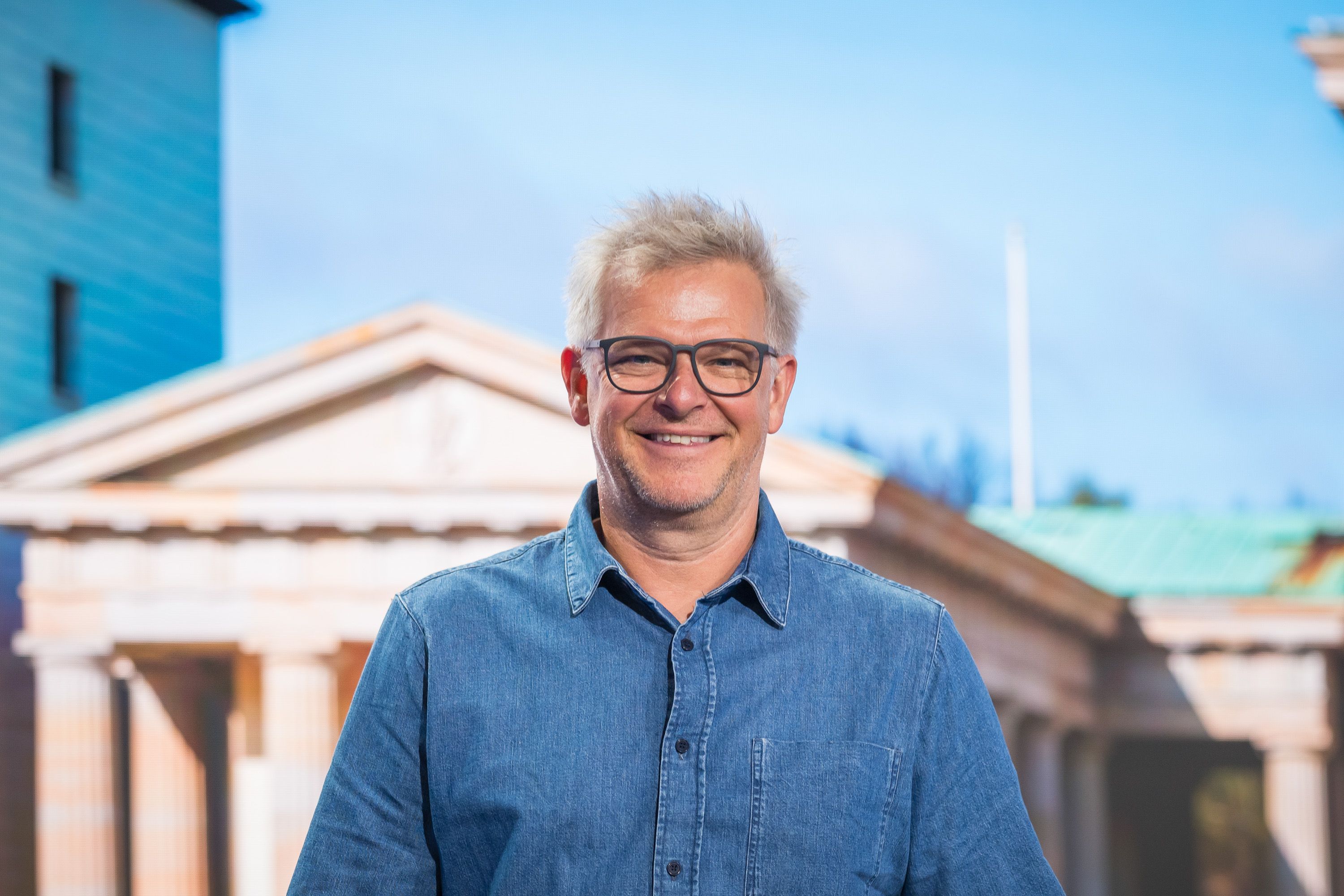 Nahaufnahme eines lächelnden Mannes mit grauen Haaren und Brille vor einem Hintergrundbild des Brandenburger Tors in Berlin.