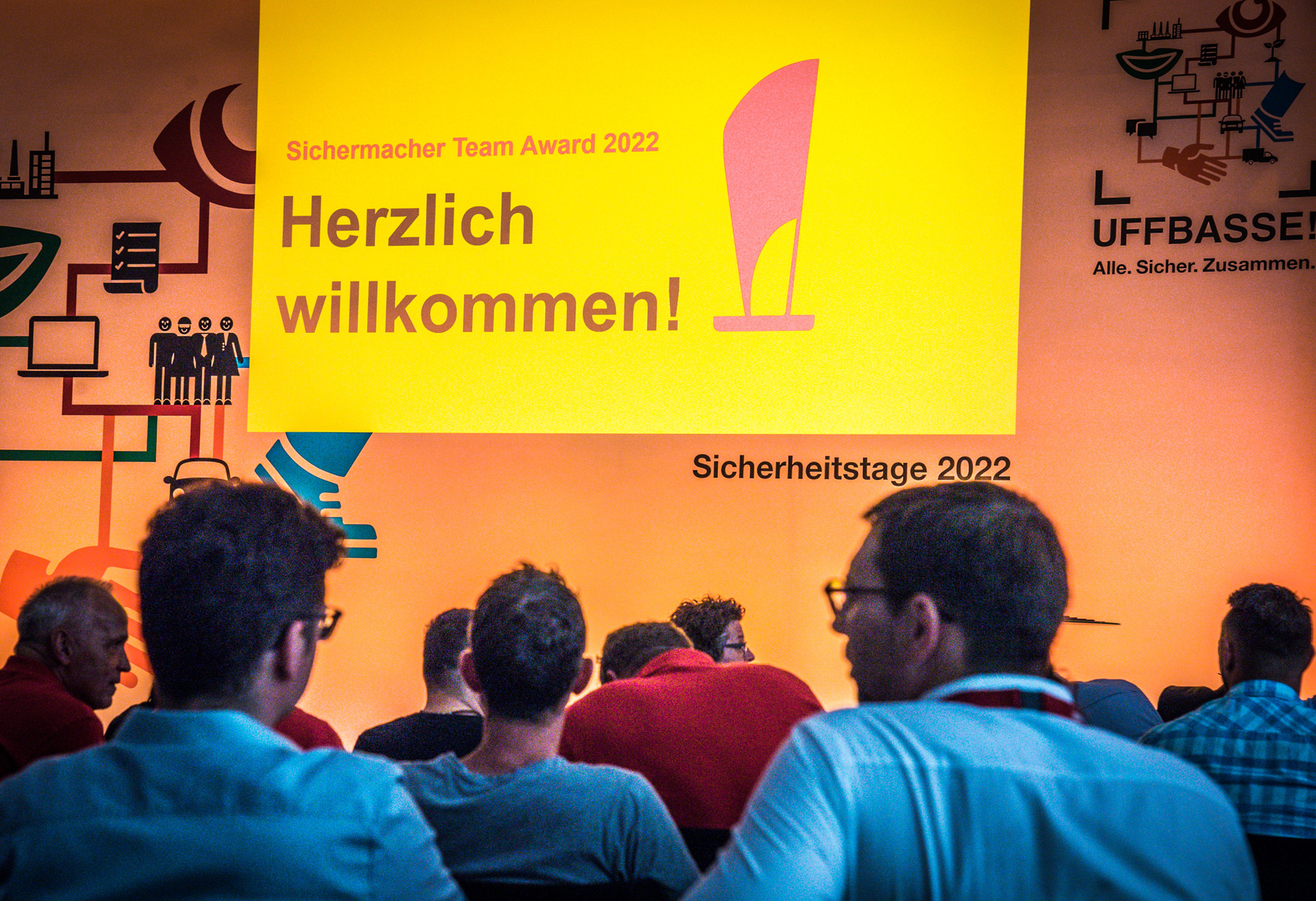 Zuschauer vor gelber Leinwand mit ‚Herzlich willkommen!‘-Folie bei einer Veranstaltung