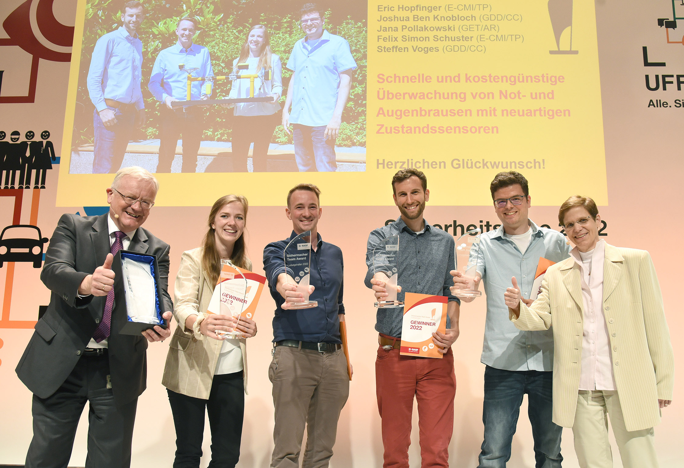Preisträger mit Urkunden auf einer Bühne vor gelber Leinwand.