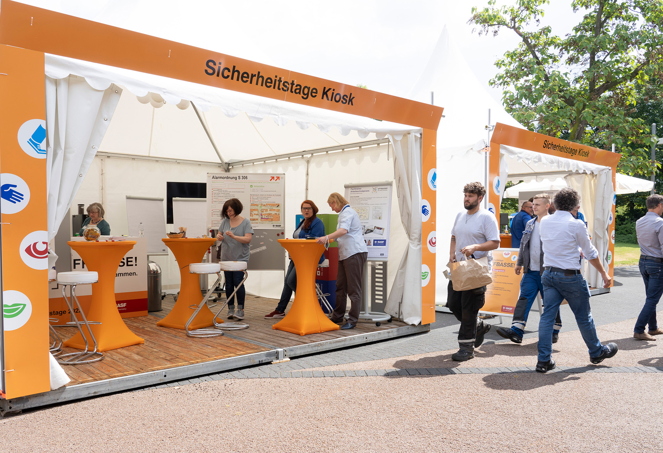 Offene Messezelte mit orangefarbener Gestaltung und Stehtischen. Personen informieren sich an den Ständen oder gehen daran vorbei.