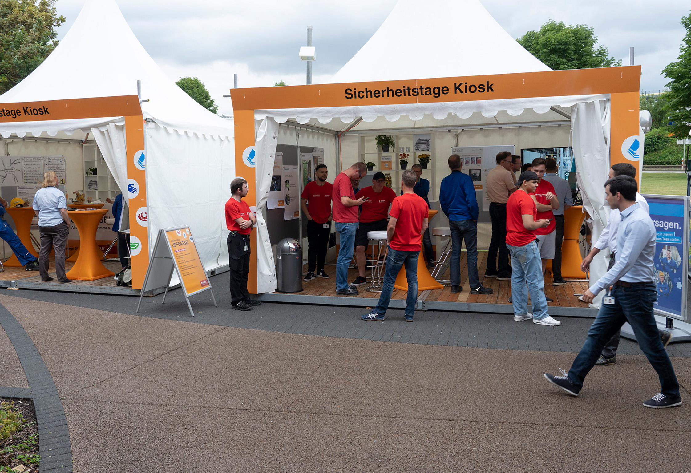 Zwei weiße Pagodenzelte mit Infoständen. Mitarbeitende in roten Shirts stehen an Stehtischen, Besucher:innen gehen am Stand vorbei.