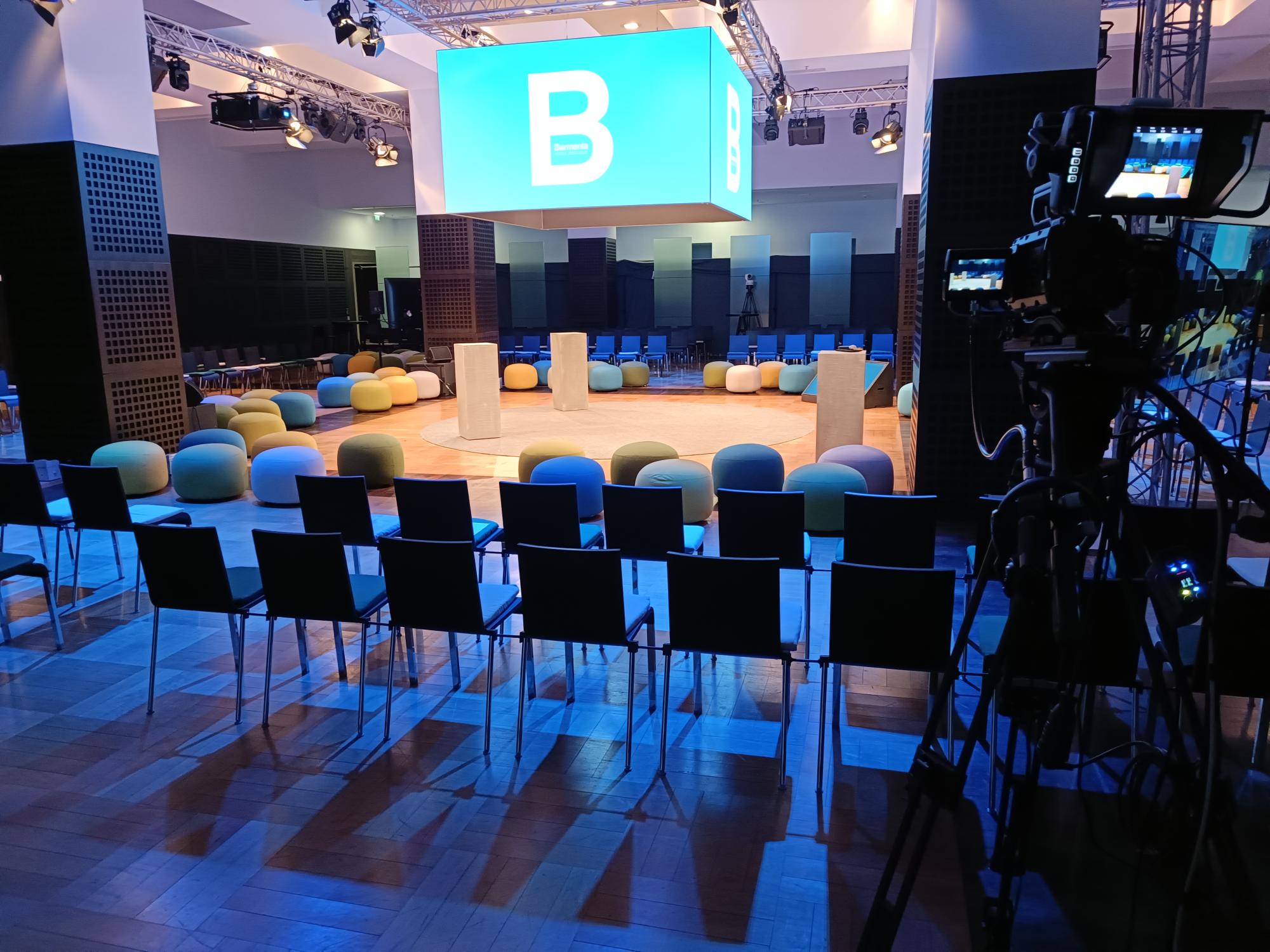 Stimmungsvoll ausgeleuchteter Veranstaltungsraum mit runder Bühne in der Mitte, umgeben von Stuhlreihen. Über der Bühne hängt ein quadratischer LED-Würfel mit einem großen Barmer-Logo.