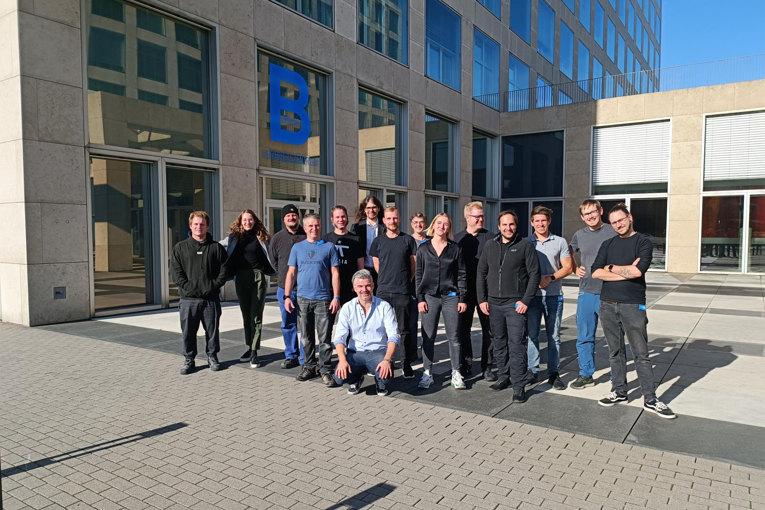 Gruppenfoto mit rund 18 Personen vor einem modernen Bürogebäude bei sonnigem Wetter. Die Gruppe steht und kniet auf einem gepflasterten Platz, alle lächeln in die Kamera