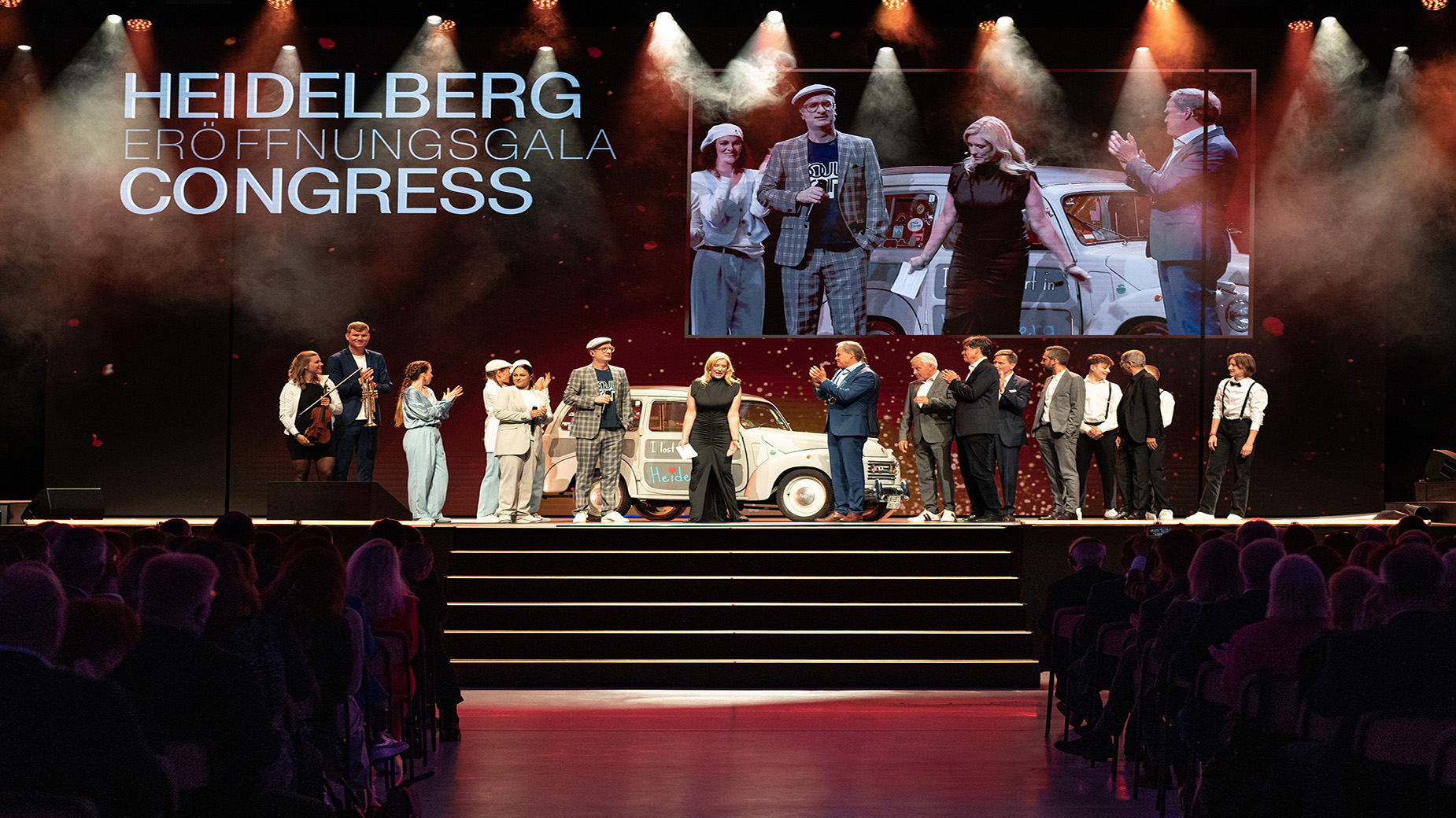 Auf einer Bühne stehen etwa 15 Personen nebeneinander. Im Hintergrund ist ein großes projiziertes Gruppenfoto zu sehen, darüber steht ‚Heidelberg Congress‘. Die Szene ist feierlich beleuchtet.