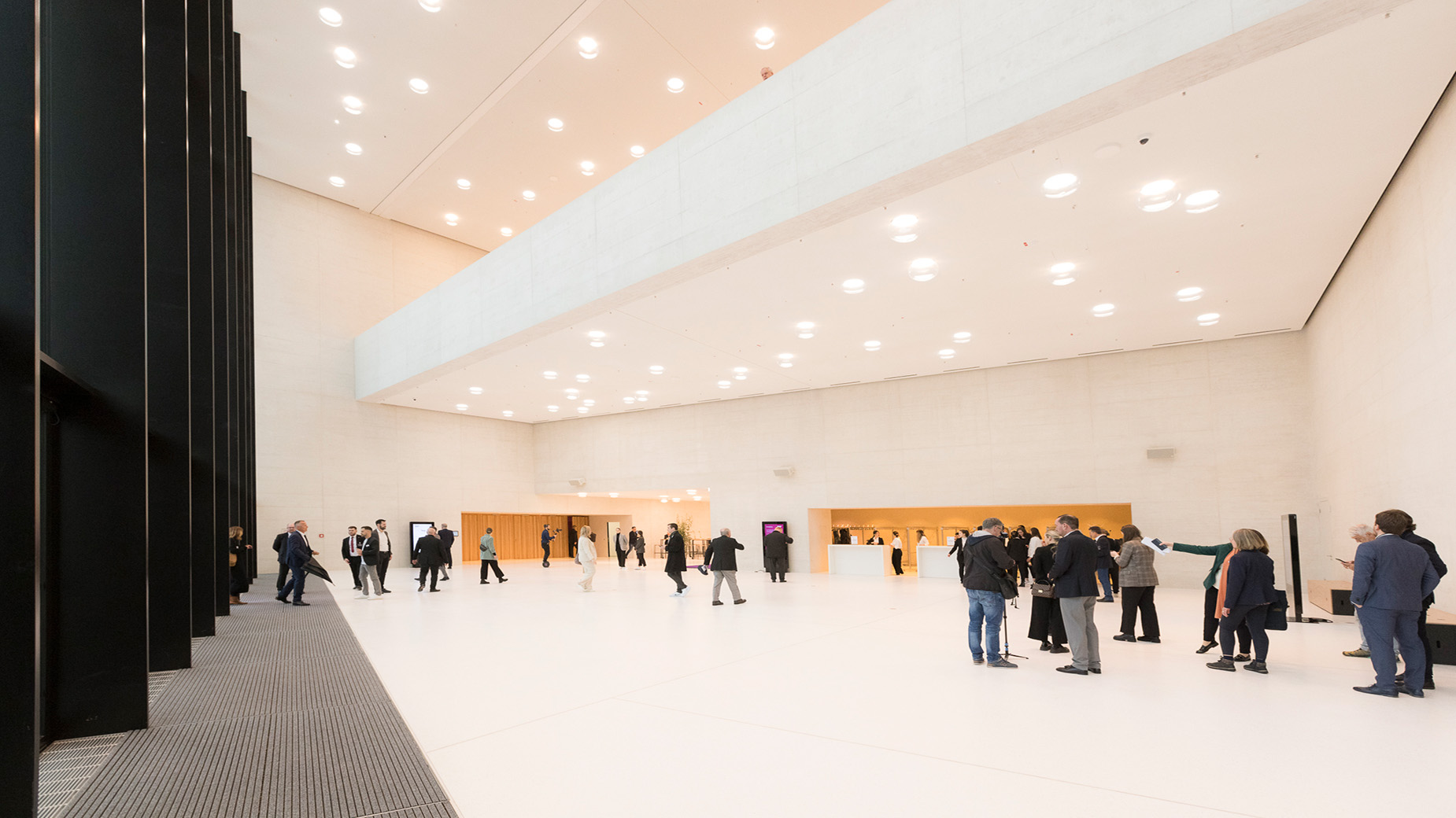 Helles und modernes HCC_Foyer mit vielen Deckenspots. Menschen in formeller Kleidung stehen in Gruppen zusammen und unterhalten sich.