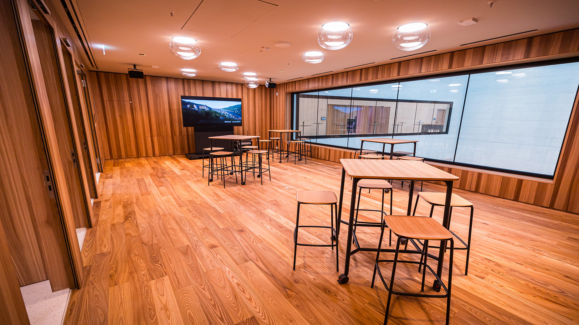 Moderner, holzverkleideter Besprechungs- oder Workshopraum mit hohen Fenstern, Stehtischen und Monitor. Der Raum wirkt warm, offen und kreativ gestaltet.