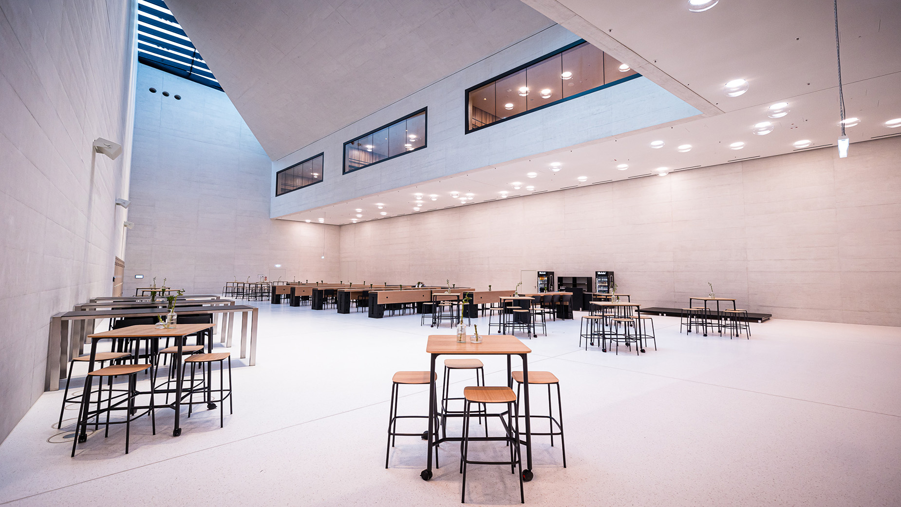 Lichter, moderner Veranstaltungsraum mit hohen Decken, weißen Wänden und Stehtischen. Die minimalistische Möblierung und die großen Fenster sorgen für eine offene, freundliche Atmosphäre.