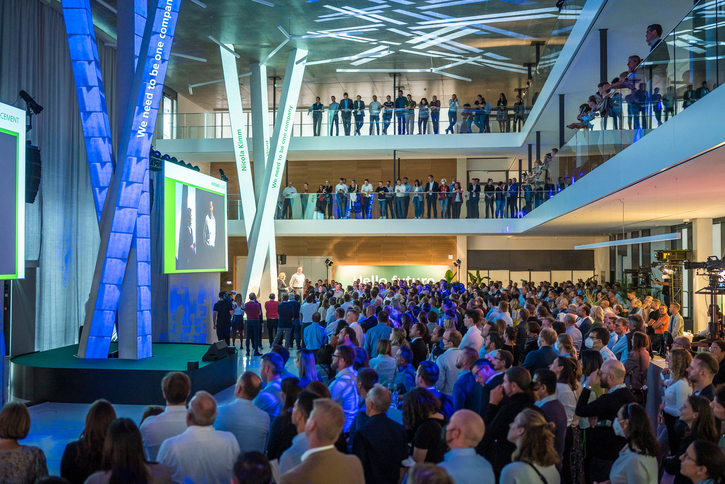 Blick in ein voll besetztes Atrium bei einer Abendveranstaltung. Auf mehreren Etagen verfolgen Gäste die Moderation auf einer großen LED-Leinwand.