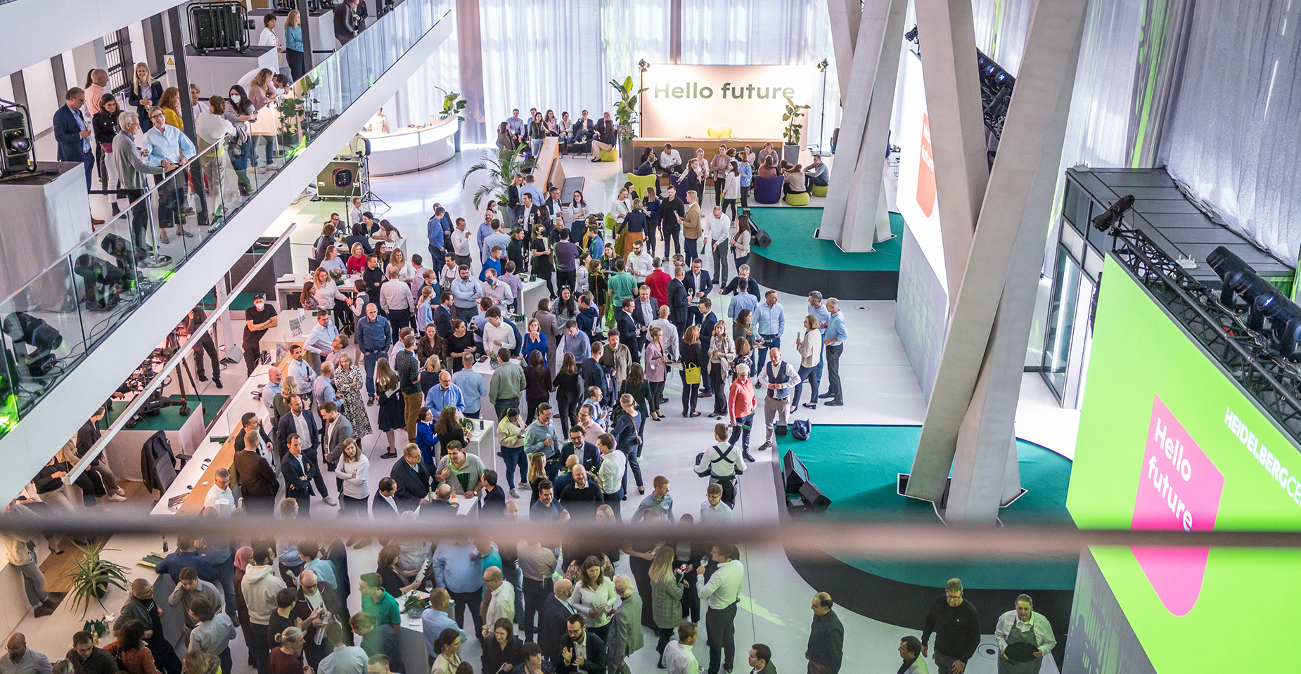Vogelperspektive auf eine Veranstaltung im Atrium. Besucher stehen in Gruppen zusammen, eine große Leinwand mit grünem Design ist gut sichtbar.