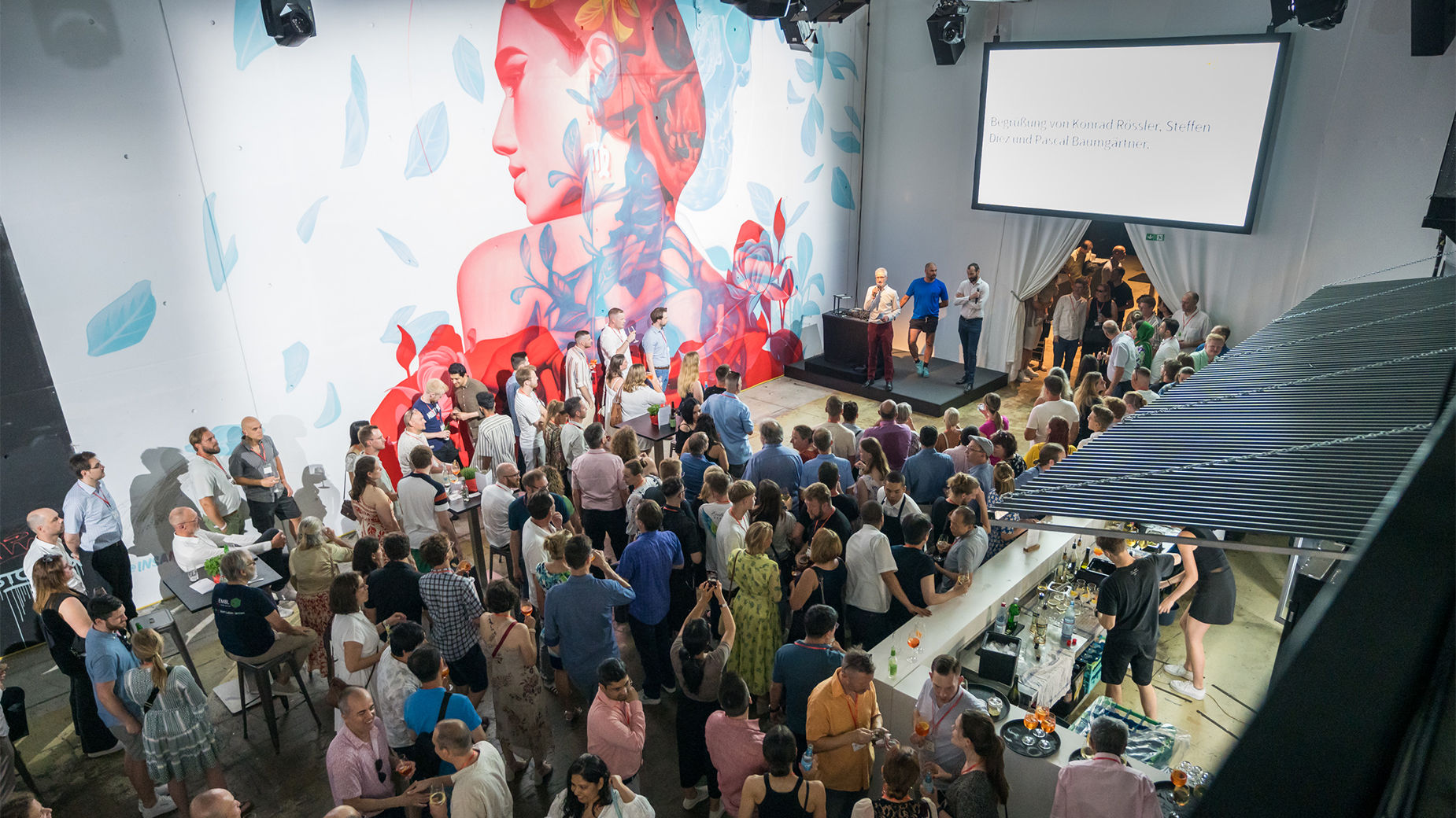 Zu sehen ist ein großes Wandgemälde, davor sind viele Gäste. Der Gastgeber begrüsst die Gäste auf einem kleinen Podium. Rechts am Bildrand befindet sich eine Bar mit Barkeepern.