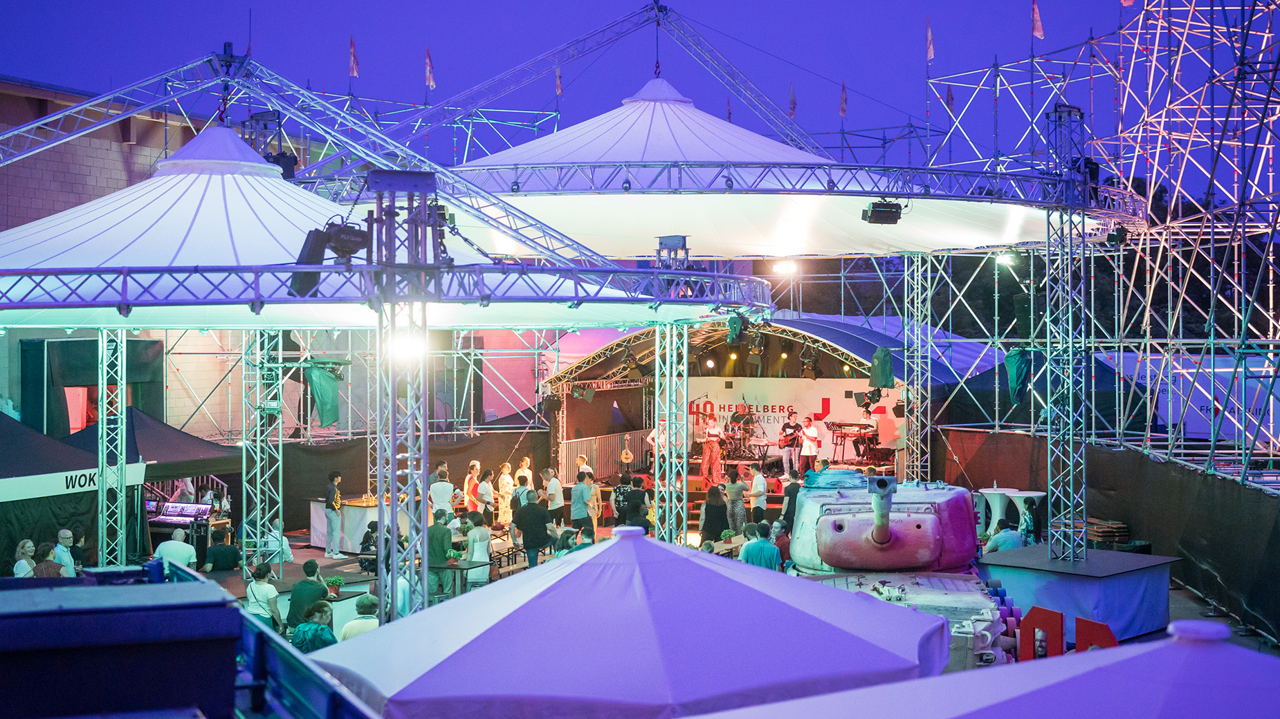 Abendliches Event mit weißen Zelten und stimmungsvoller Beleuchtung. Menschen sitzen an gedeckten Tischen in einem festlich dekorierten Außenbereich.