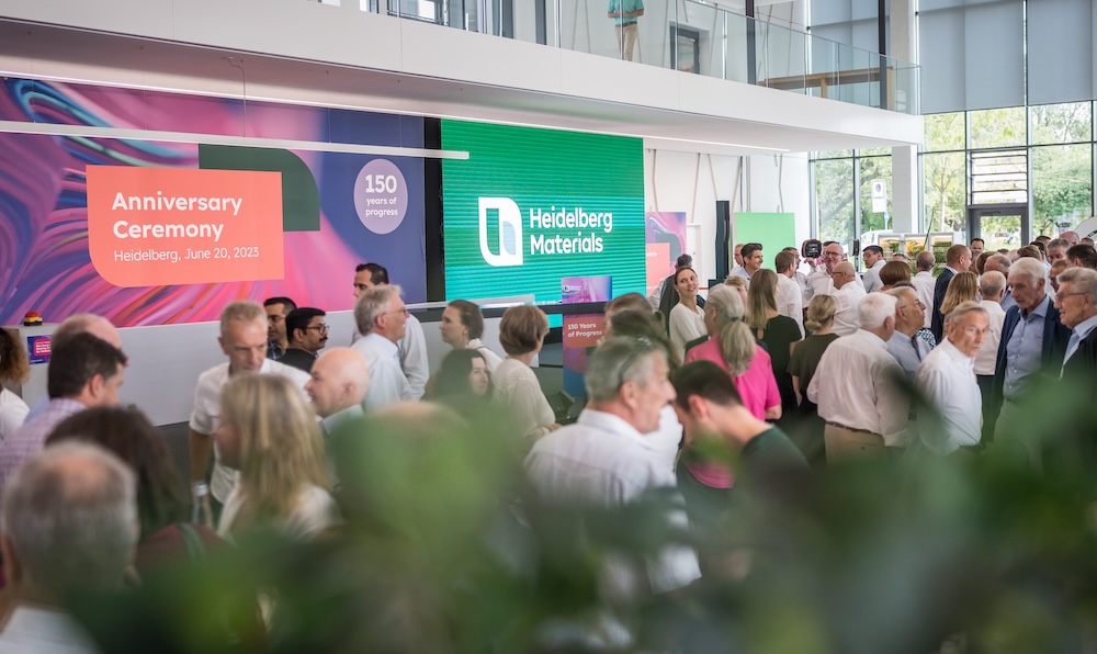 Großes Get-together bei einer Jubiläumsfeier von Heidelberg Materials mit zahlreichen Gästen in Businesskleidung in einem modernen, lichtdurchfluteten Gebäude. Auf den digitalen Bildschirmen ist das Key Visual der Veranstaltung zu sehen.