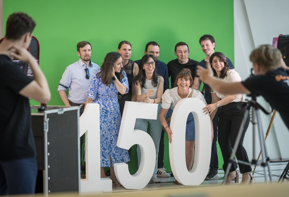 Eine fröhliche Gruppe von zehn jungen Erwachsenen posiert vor einem grünen Hintergrund mit großen weißen Zahlen „150“, die auf das Jubiläum hinweisen. Zwei Personen am Bildrand fotografieren die Szene.