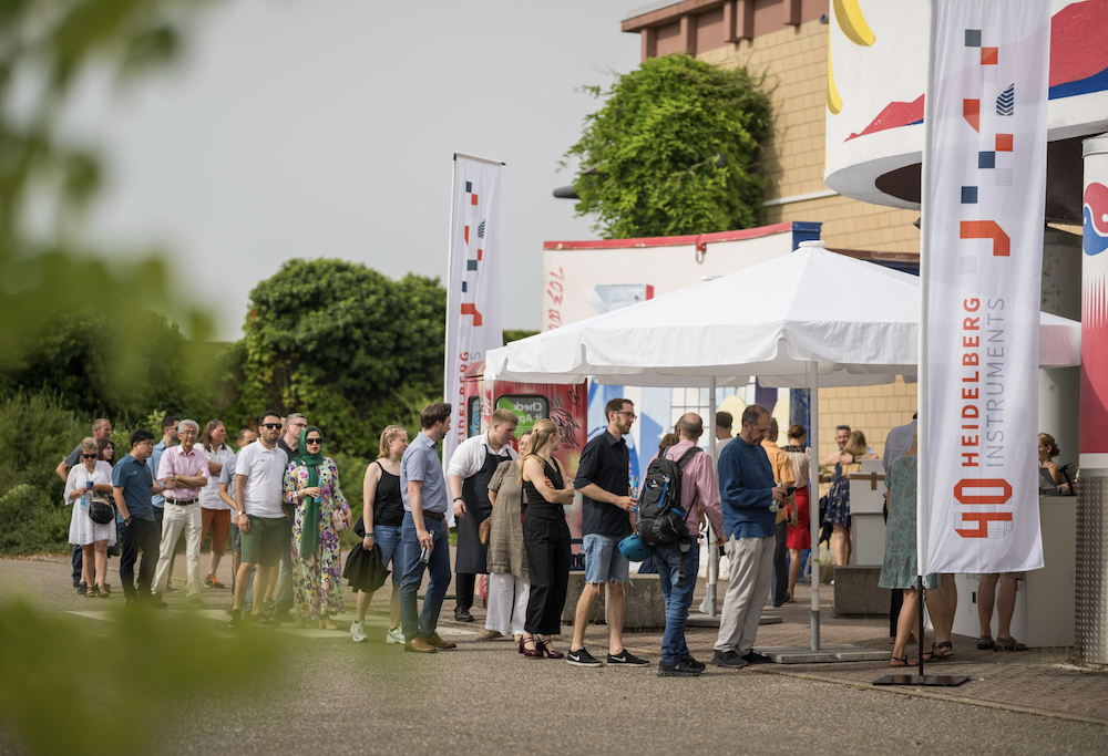 Menschen stehen bei sonnigem Wetter in einer Schlange vor einem weißen Pavillon mit dem Logo „Heidelberg Instruments“ und der Zahl „40“ auf einem Banner. Die Veranstaltung findet im Freien vor einem farbenfrohen Gebäude statt.
