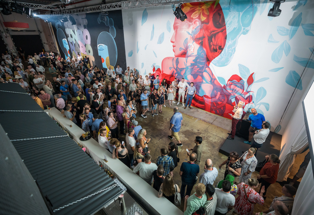 Große Gruppe von Menschen steht in einer Halle vor einer farbenfrohen Wand mit zwei großformatigen Wandgemälden – eines zeigt ein stilisiertes Frauenporträt mit floralen Elementen. Eine Person spricht auf einer kleinen Bühne.