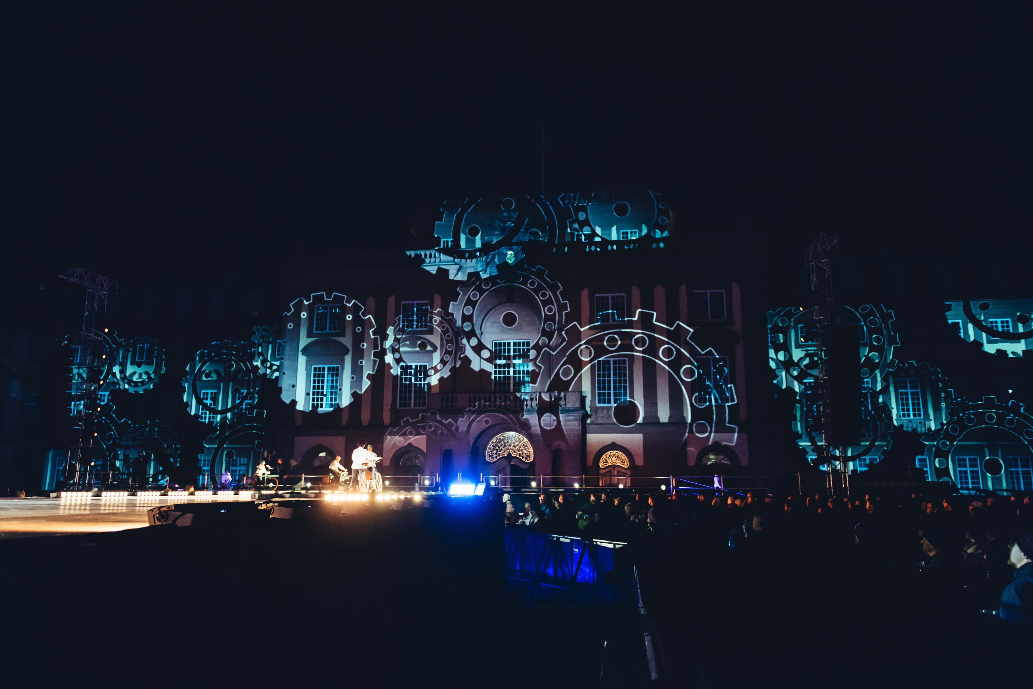 Nächtliche Lichtshow auf der Fassade eines historischen Gebäudes, auf das ein leuchtendes Fahrrad und animierte Lichtmuster projiziert werden. Viele Zuschauer*innen stehen im dunklen Außenbereich und filmen das Spektakel mit ihren Smartphones.