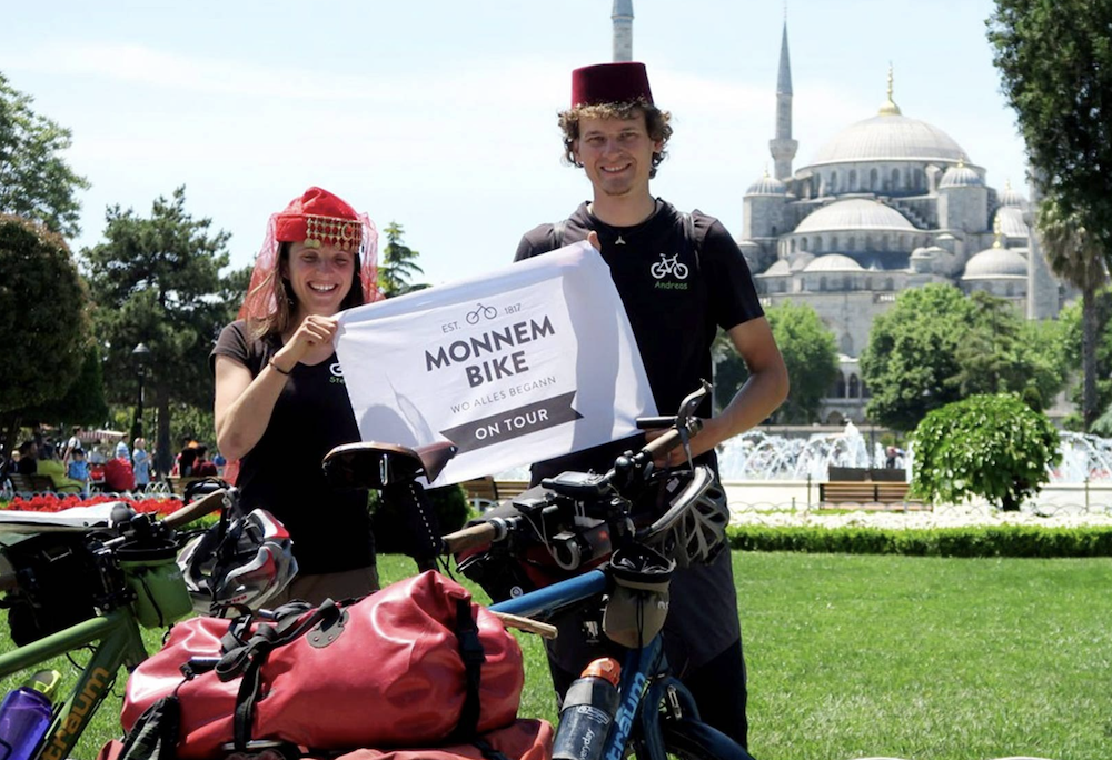 Zwei lächelnde Radreisende mit traditionellen roten Hüten halten ein Schild mit der Aufschrift „Monnem Bike – Wo alles begann – On Tour“. Vor ihnen stehen bepackte Fahrräder, im Hintergrund ist die Blaue Moschee in Istanbul in einem Park zu sehen.