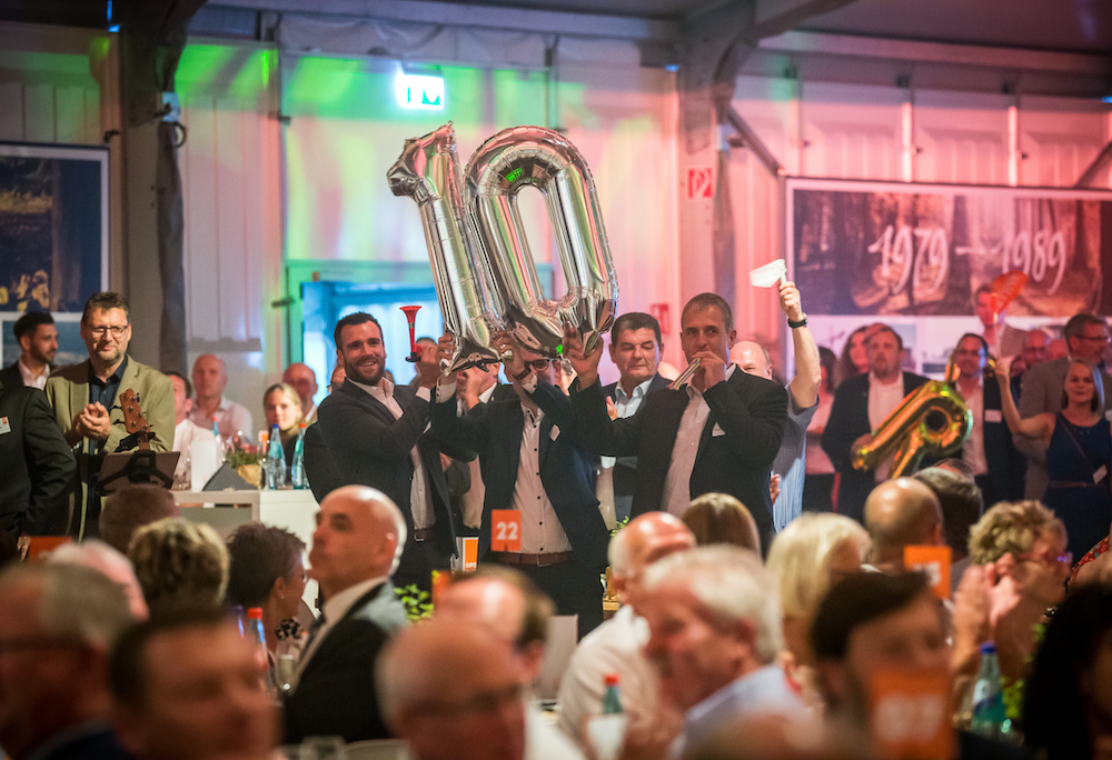 Männer in festlicher Kleidung heben bei einer Veranstaltung silberne Ballons mit der Zahl „10“ in die Höhe. Die Feier findet in einem festlich beleuchteten Saal mit vielen sitzenden Gästen statt. Im Hintergrund feiern weitere Personen.