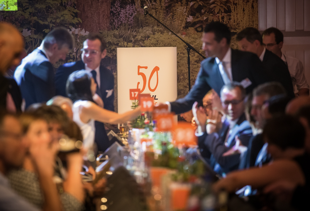 Abendliche Jubiläumsfeier mit festlich gekleideten Gästen an einer langen, dekorierten Tafel. Im Zentrum steht ein beleuchtetes Schild mit der Aufschrift „50“. Personen begrüßen sich per Handschlag, die Stimmung ist feierlich und lebhaft.