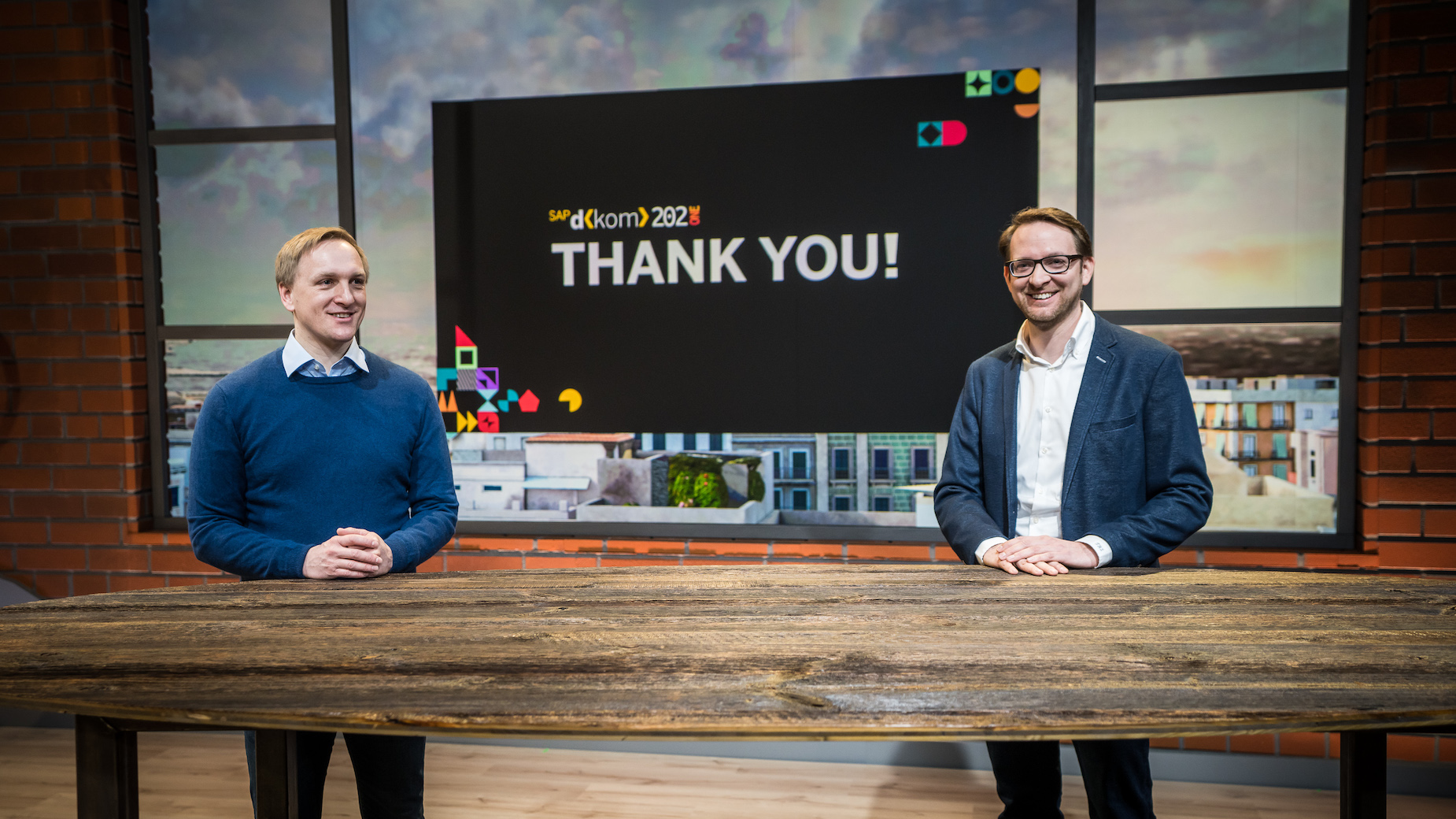 Zwei Männer stehen in einem virtuellen Studio vor einer Leinwand mit der Aufschrift ‚Thank you‘. Der Raum ist modern inszeniert mit digitalen Elementen.