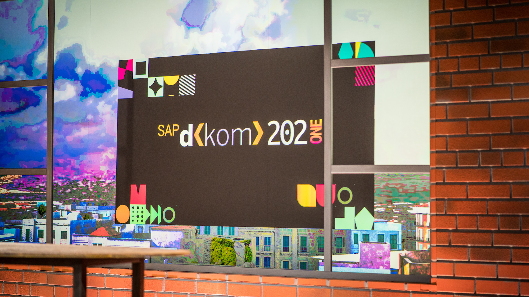 Digitales Bühnenbild mit dem Logo ‚SAP d-kom 2021‘. Die Gestaltung zeigt virtuelle Fensterfronten, Stadtgrafik und Post-its – tech-inspirierte Kulisse