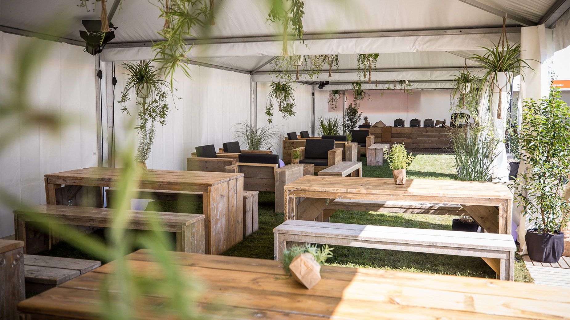 Helles Zelt mit rustikalen Holzmöbeln, grüner Rasenfläche und hängenden Pflanzen – gemütliche Eventlounge im Grünen.