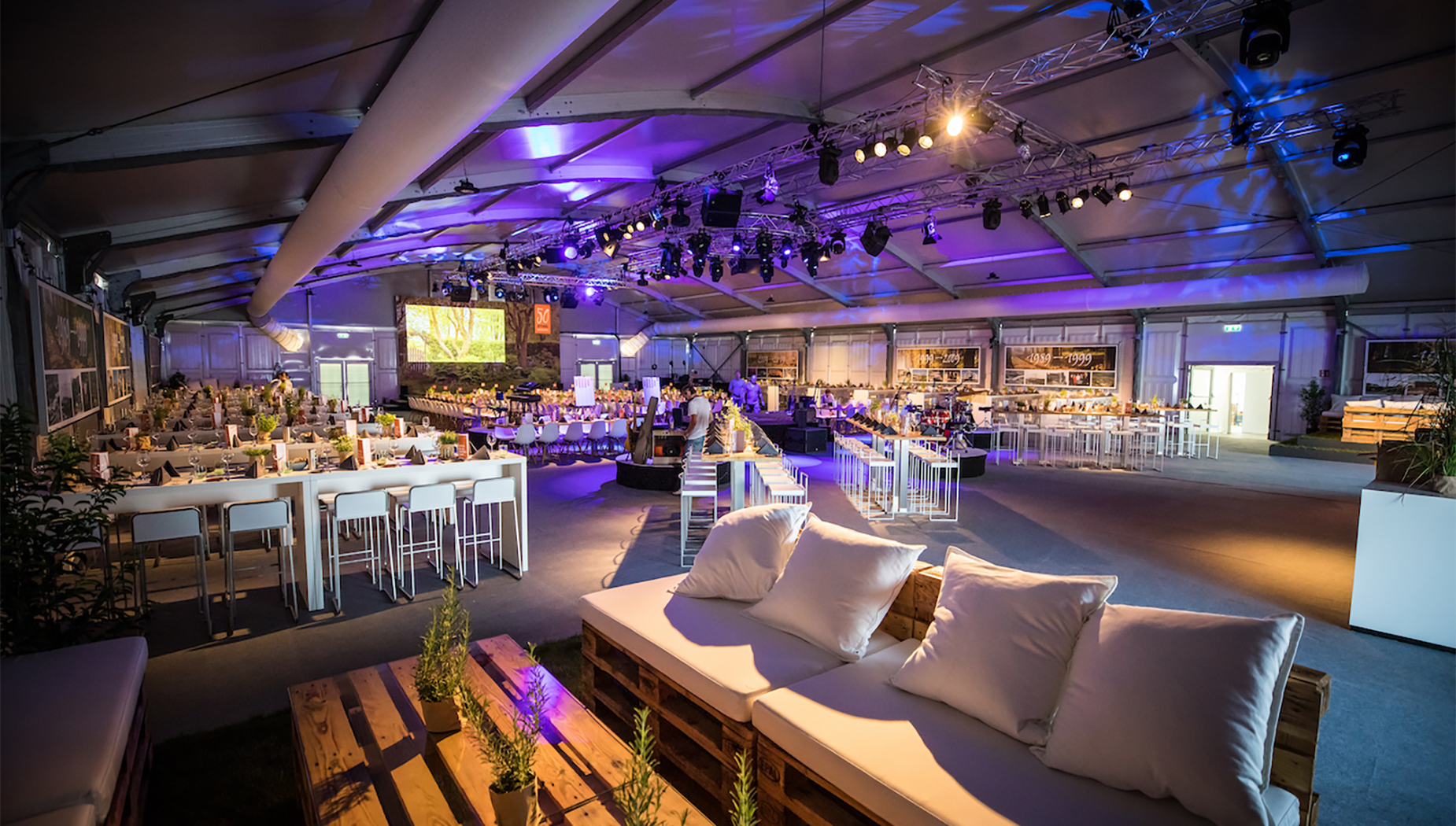 Stimmungsvoll beleuchtete Eventhalle mit festlich gedeckten Tischen, Lounge-Ecken und großer Bühne zum Firmenjubiläum.