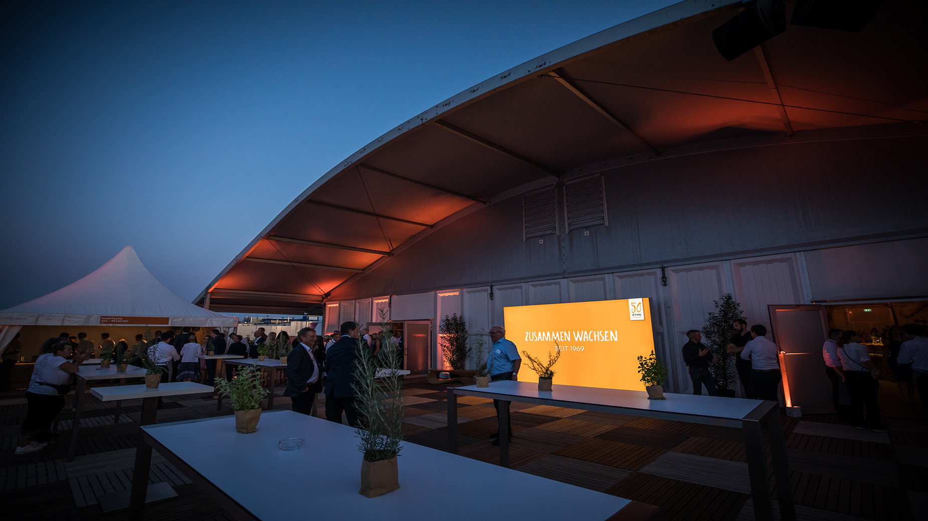 Beleuchteter Outdoorbereich am Abend mit oranger Wand „Zusammen wachsen – seit 1969“.