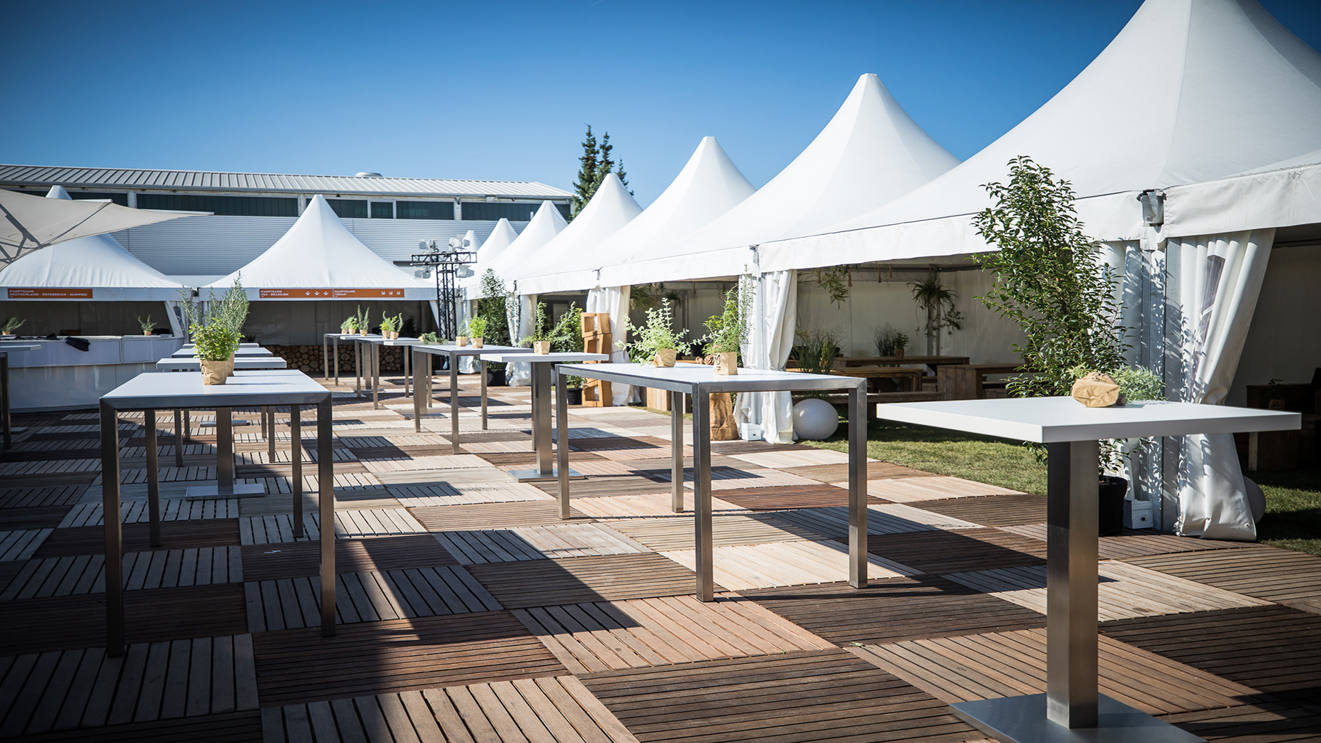 Reihe moderner Stehtische mit Kräuterdeko vor weißen Pagodenzelten bei Sonnenschein.