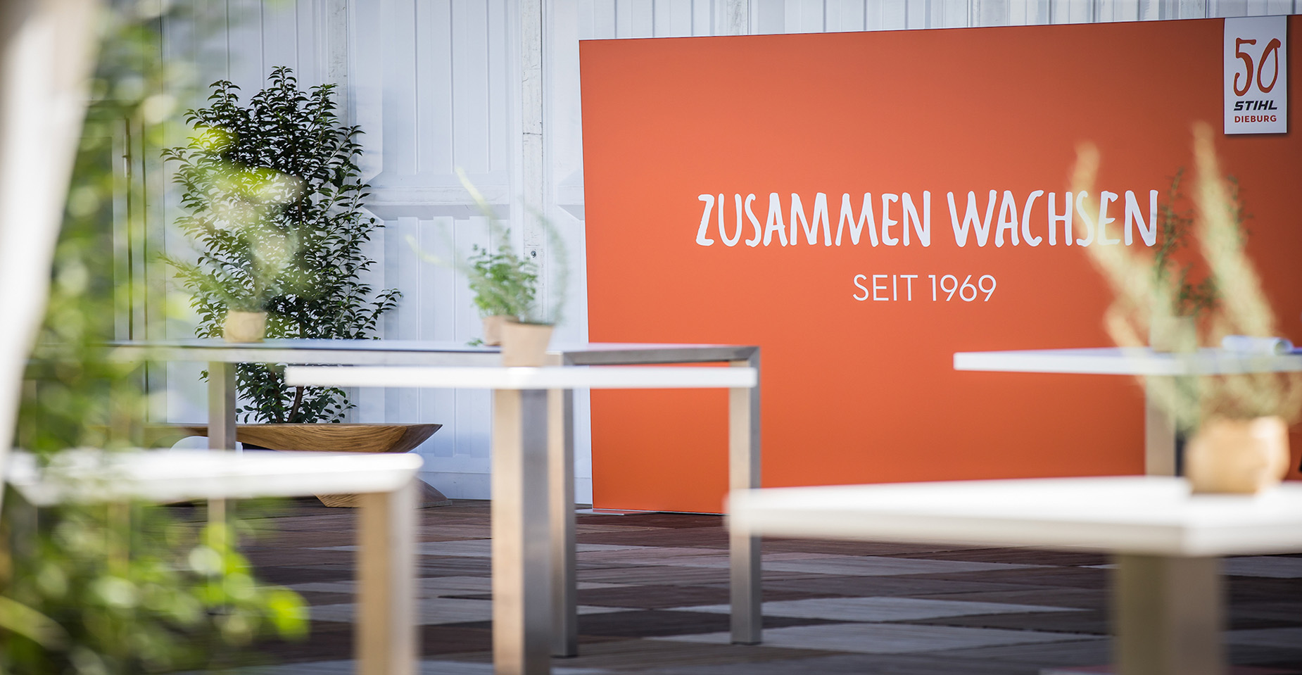 Orangene Wand mit dem Jubiläumsslogan „Zusammen wachsen – seit 1969“ und modern dekorierter Eventfläche mit Pflanzen und Stehtischen.