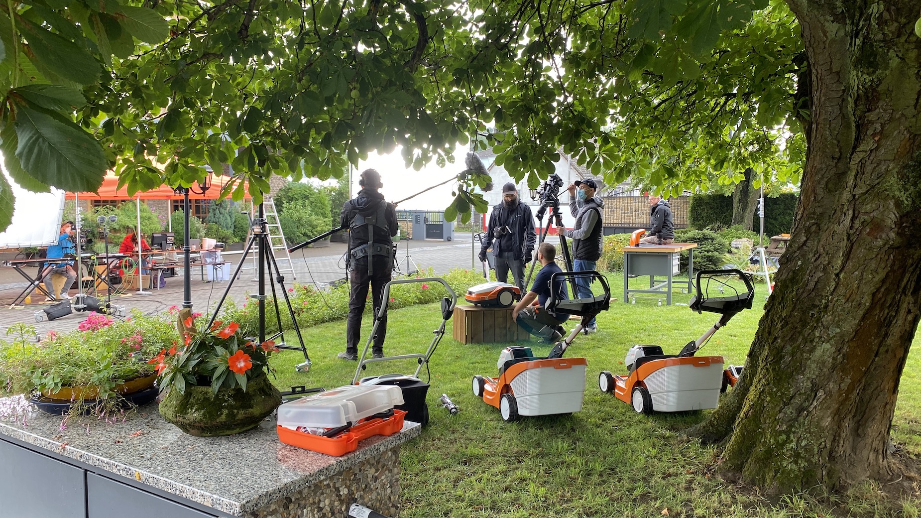Außendreh unter Bäumen mit Technikcrew und Kabelkisten. Im Vordergrund stehen Transportkoffer, im Hintergrund Menschen an Kamera- und Tonpositionen.