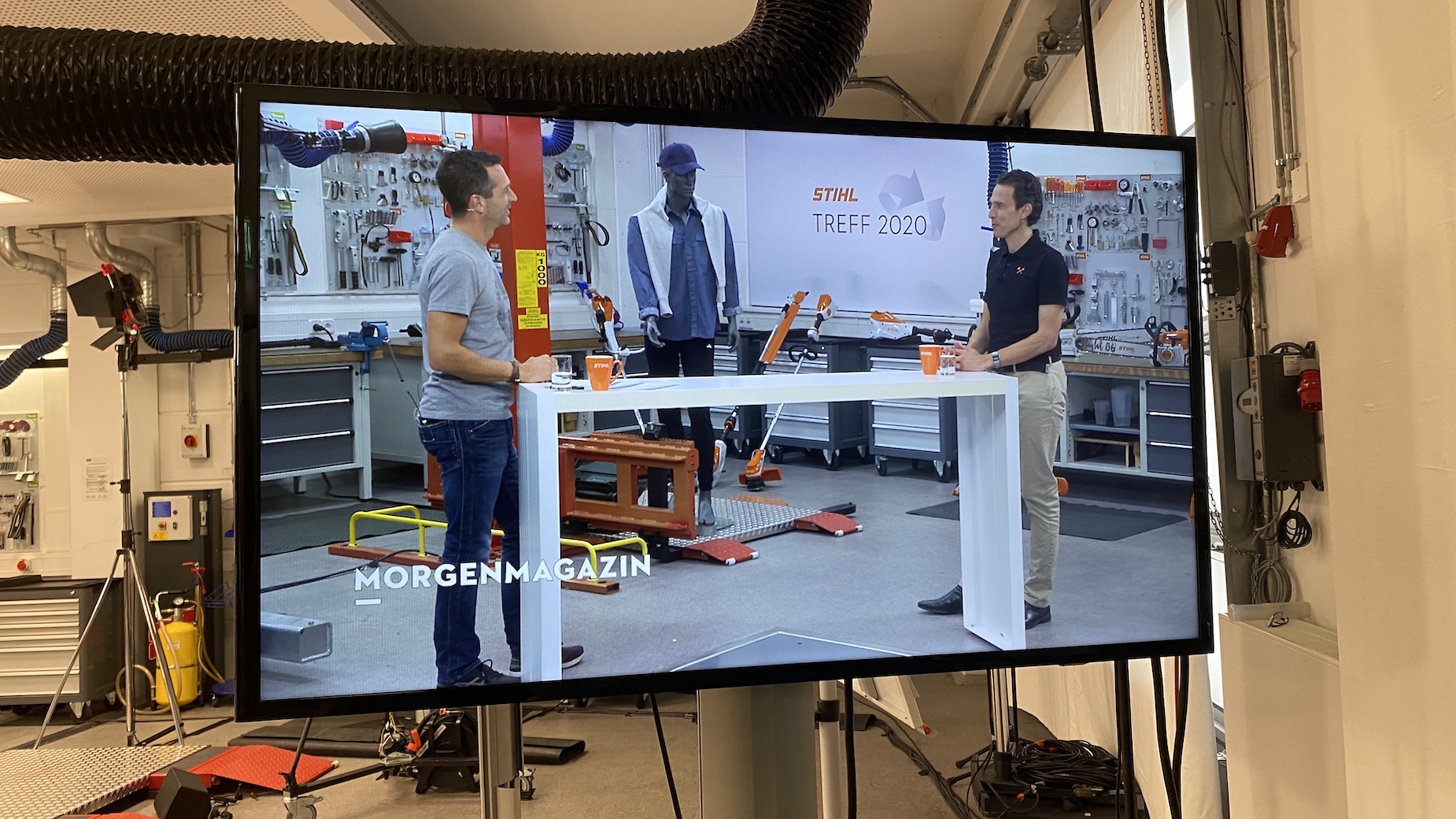 Kameraaufnahme in einem Labor- oder Technikstudio mit zwei Personen vor einem Arbeitstisch, ein Kameramonitor zeigt die Szene live