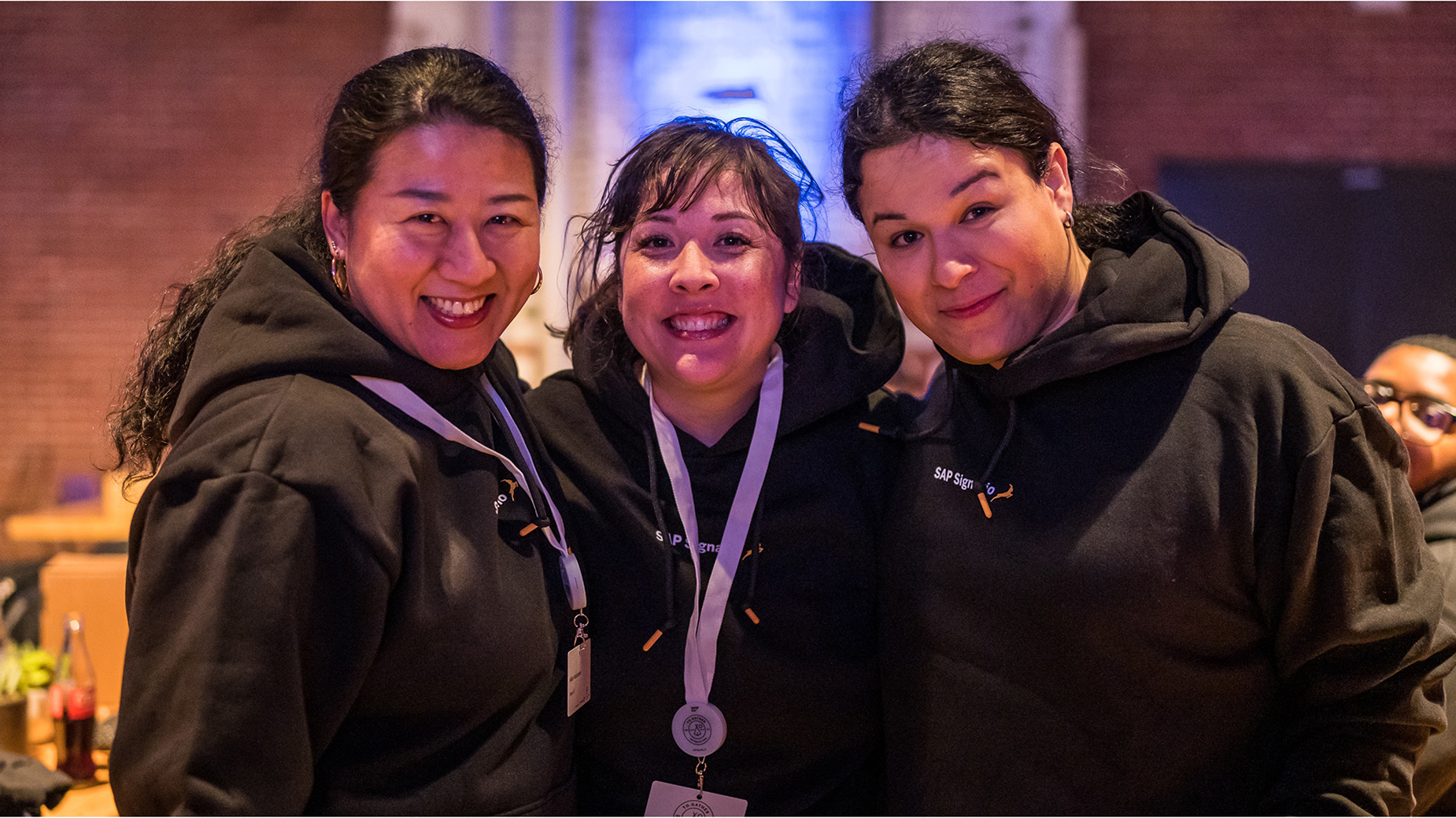 Drei Frauen in schwarzen Hoodies lächeln in die Kamera, eine trägt eine Medaille um den Hals.