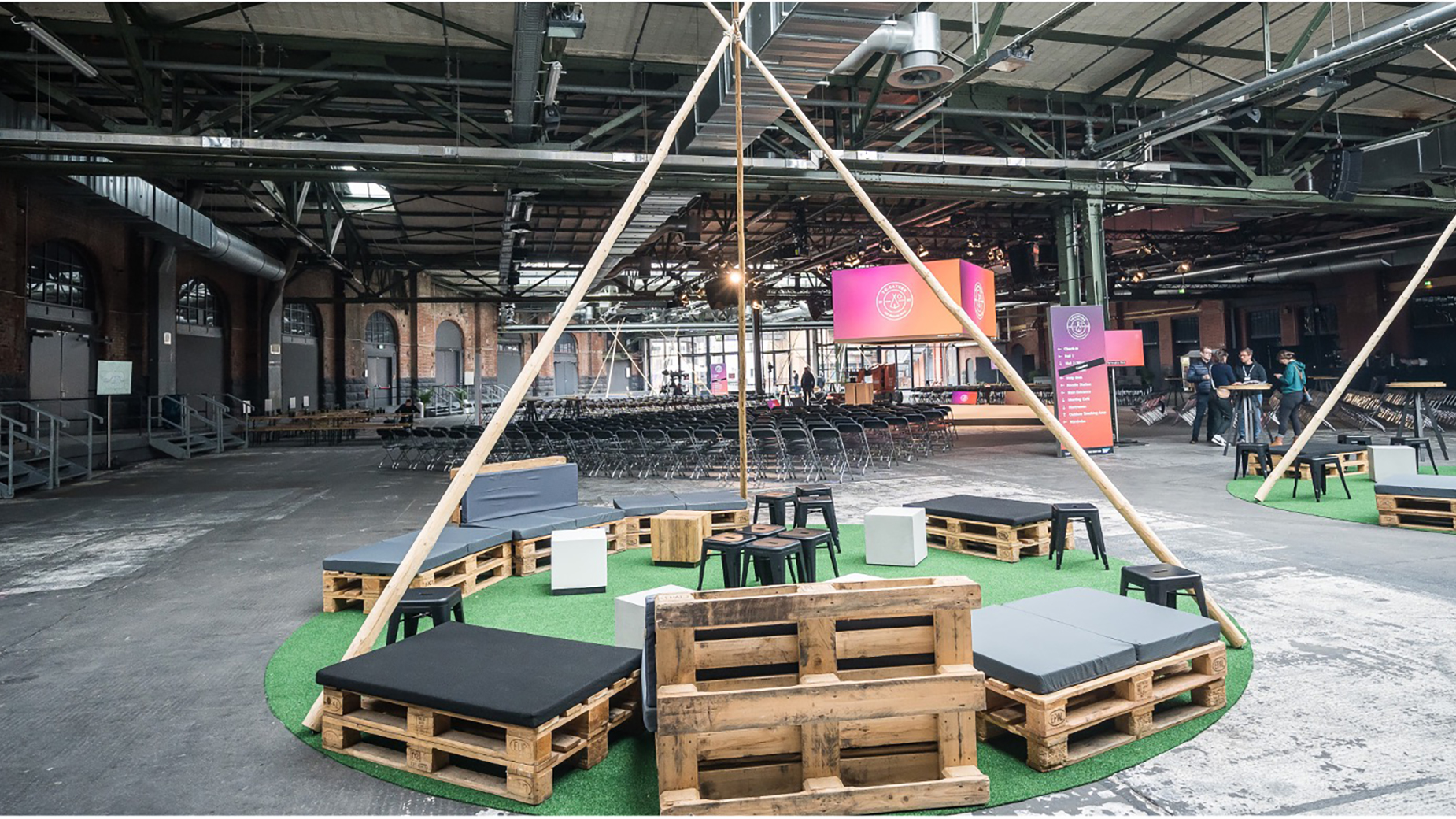 Lounge-Bereich mit Palettenmöbeln und grünem Teppich in industrieller Umgebung. Im Hintergrund hängt ein Banner mit dem Logo der Veranstaltung.