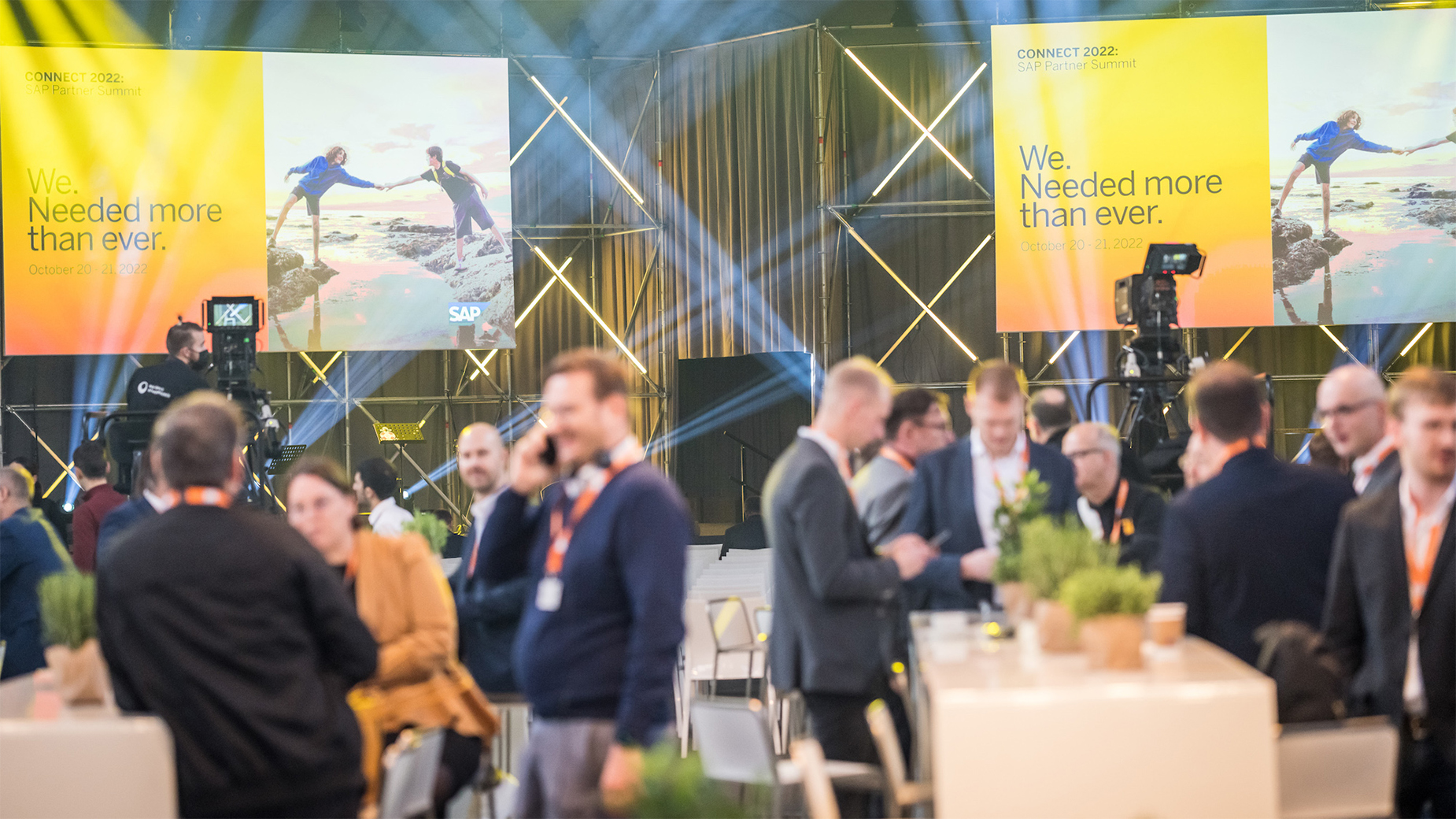 Im Hintergrund befindet sich die Hauptbühne der CONNECT. Im Vordergrund stehen und sitzen die Besucher*innen an den Tischen.