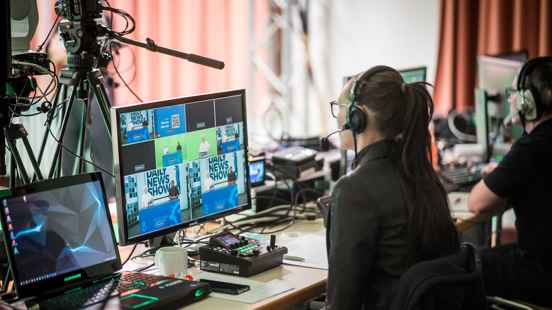 Zwei Personen mit Headsets arbeiten in der Regie. Auf Monitoren sind verschiedene Kameraperspektiven einer Talkshow mit Greenscreen zu sehen.