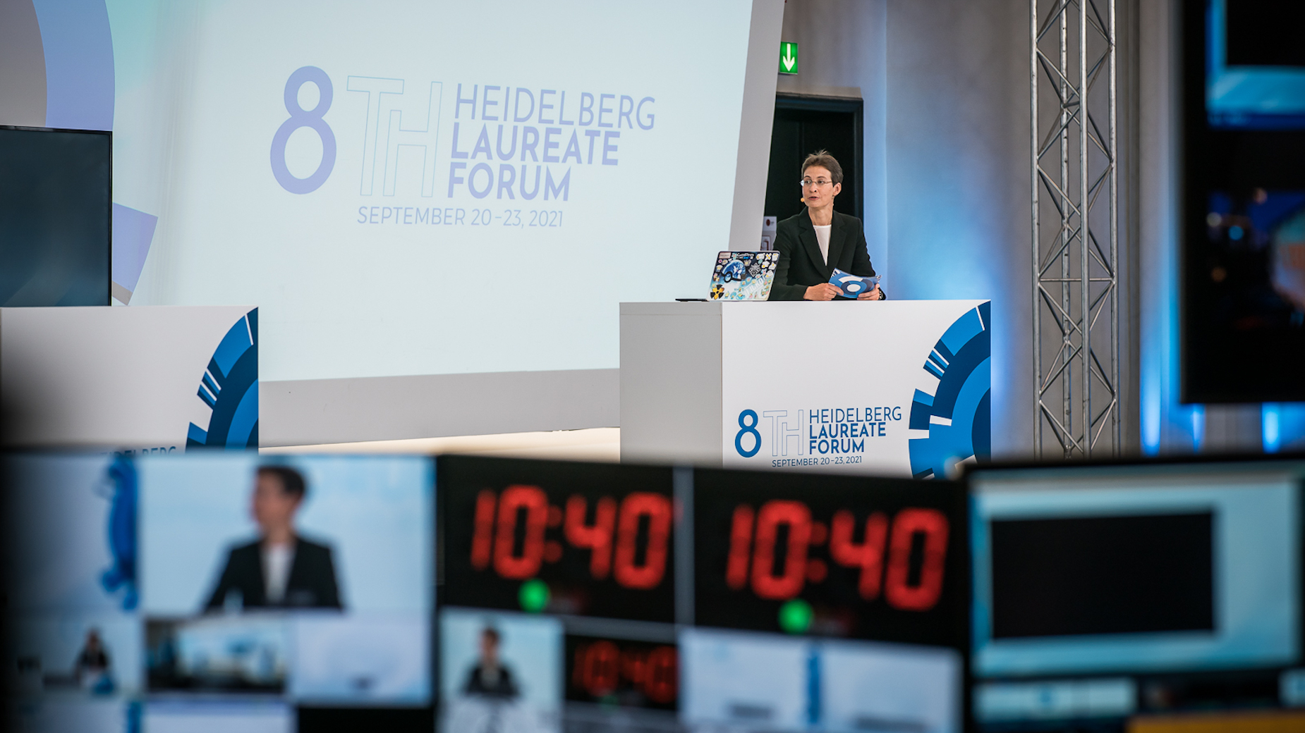 Moderatorin spricht auf der Bühne des 8th Heidelberg Laureate Forum. Im Vordergrund ist die Regie mit mehreren Timern und Monitoren zu sehen.
