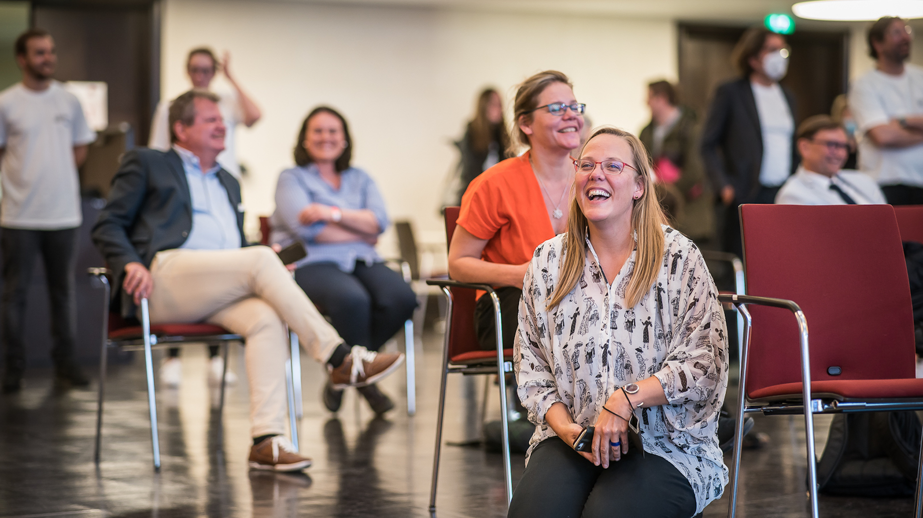 Mehrere Personen sitzen lachend in einem Veranstaltungsraum und folgen aufmerksam dem Geschehen.