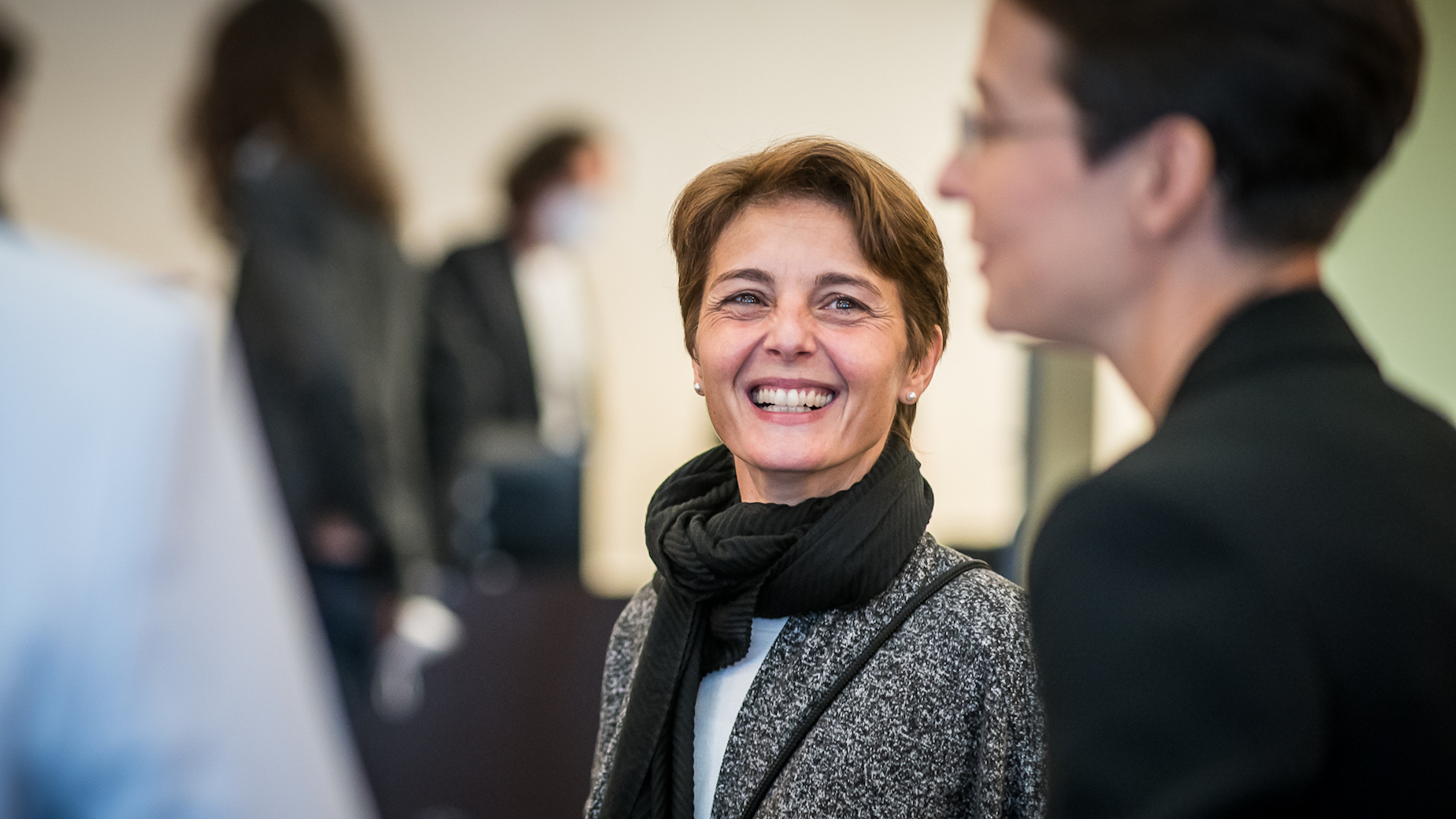 Frau mit kurzem Haar und grauem Schal lächelt in die Kamera, während sie sich mit einer anderen Person unterhält.