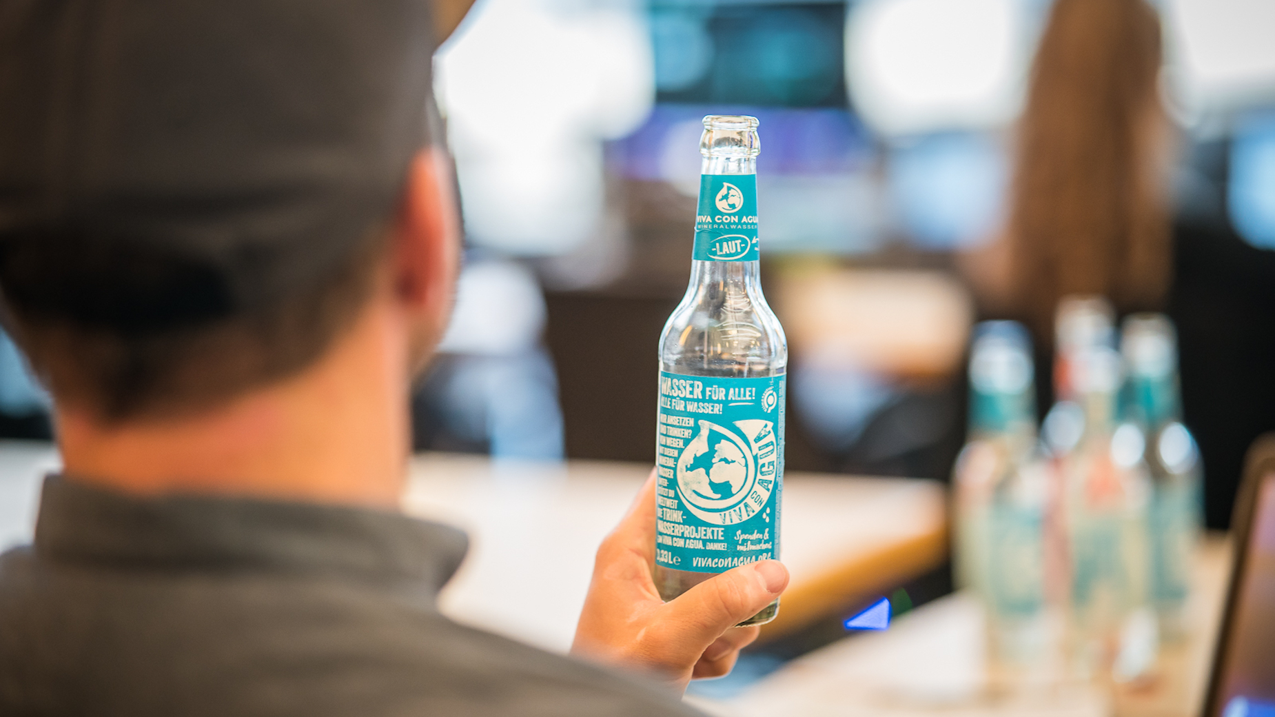 Person hält eine Flasche ‚Viva con Agua‘ mit türkisfarbenem Etikett in der Hand. Im Hintergrund verschwimmen Menschen und Bildschirme.