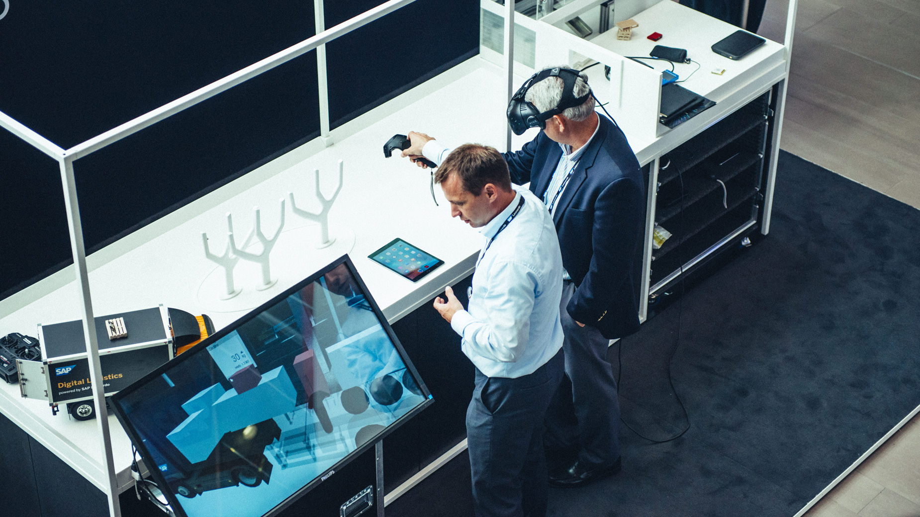 Zwei Männer testen interaktive VR-Anwendungen an einem Messestand mit Touchscreen.