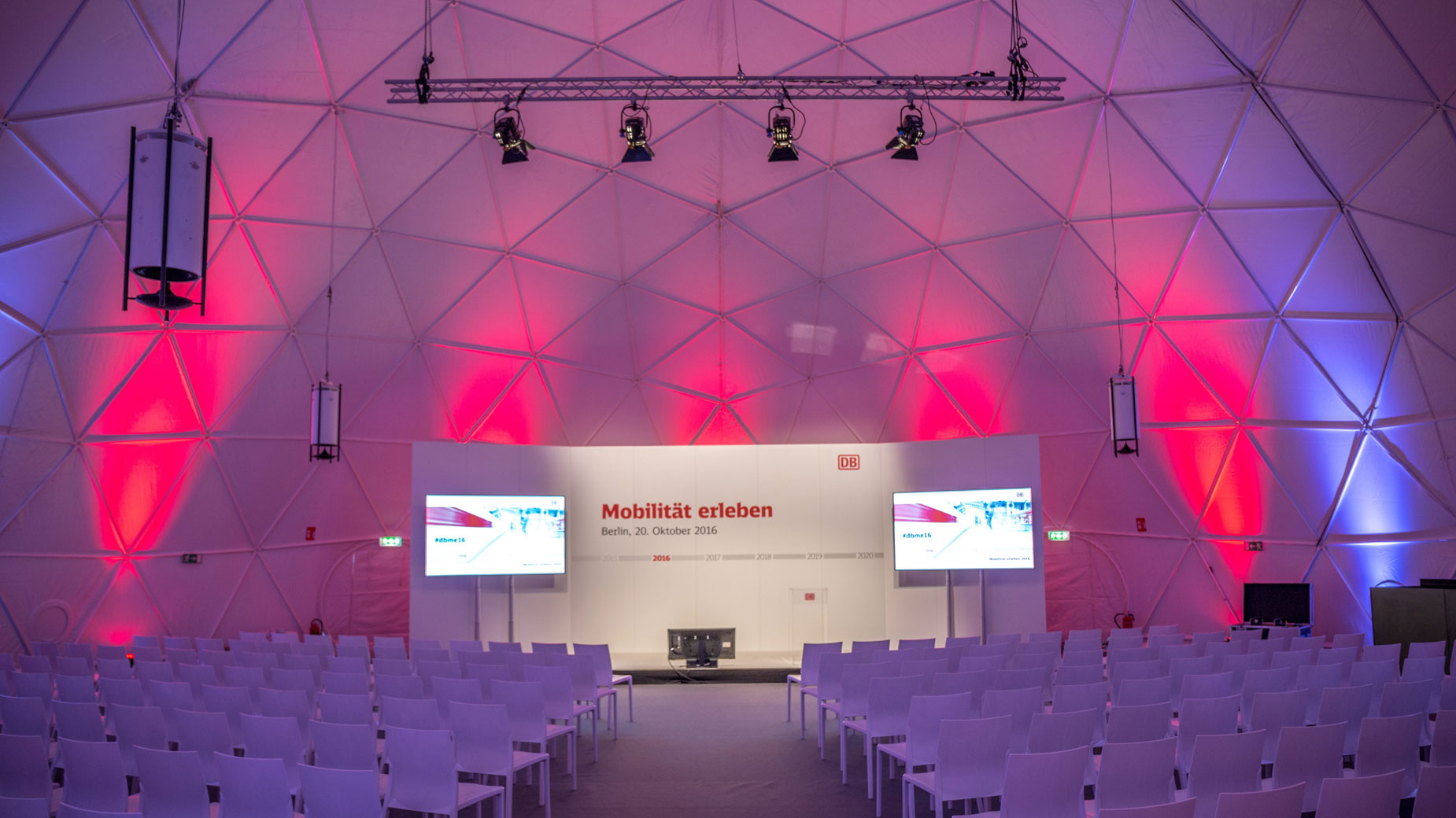 Beleuchtete Eventlocation mit Bühne, Leinwänden und Sitzreihen – bereit für eine Präsentation oder Konferenz.
