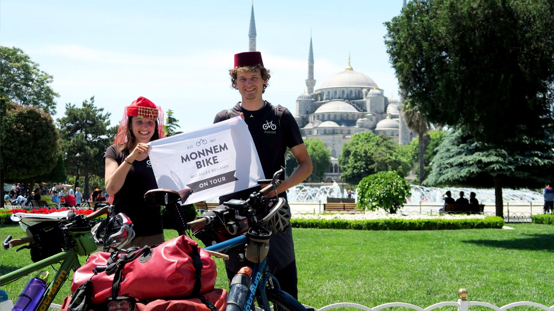 Zwei Fahrradreisende posieren mit Monnem-Bike-Schild vor dem historischen Hintergrund von Istanbul.