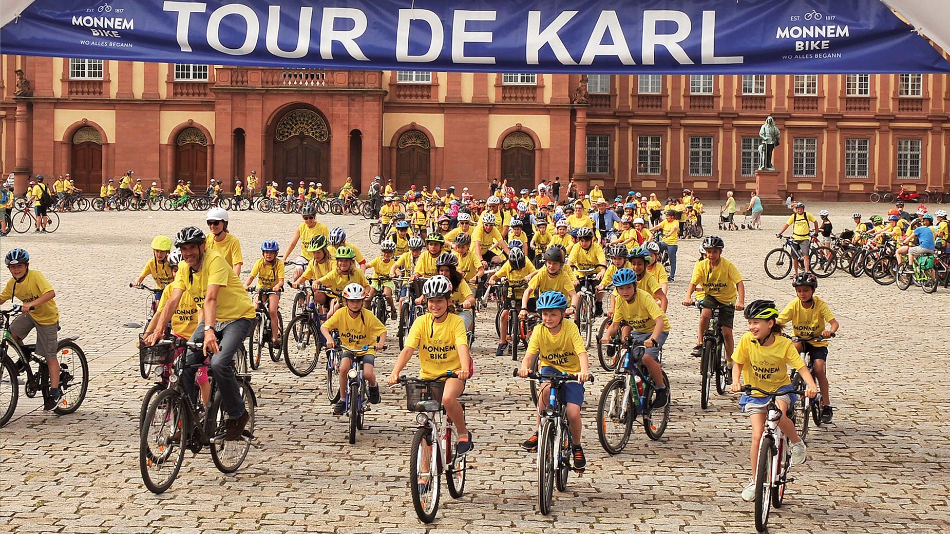 Fahrradgruppe in gelben T-Shirts startet zur Aktion ‚Tour de Karl‘ vor dem Barockschloss Mannheim.