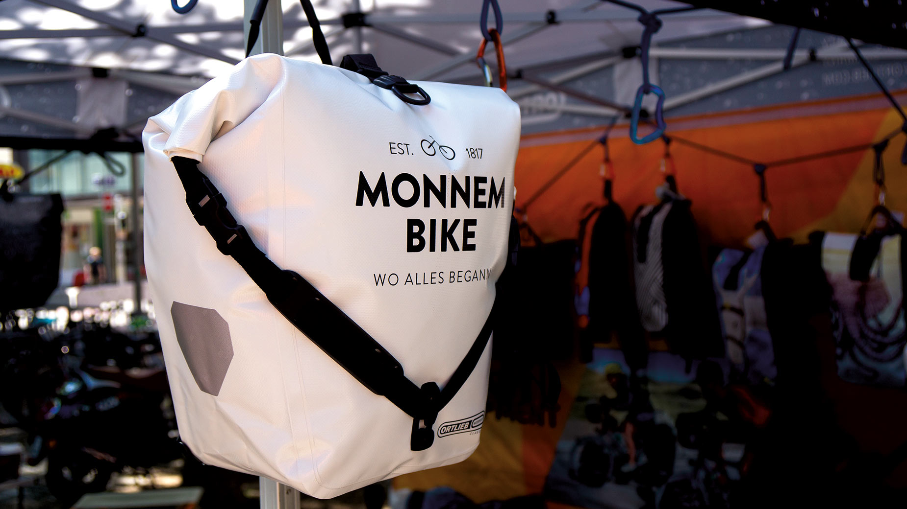 Weiße Fahrradtasche mit Monnem-Bike-Aufdruck hängt an einem Fahrrad auf einem Eventgelände.