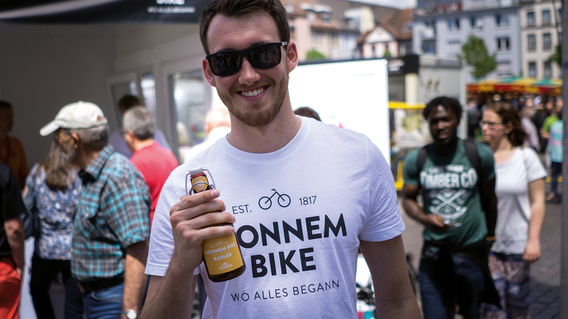Mann mit Sonnenbrille und Monnem-Bike-T-Shirt hält ein Getränk auf einer Veranstaltung.